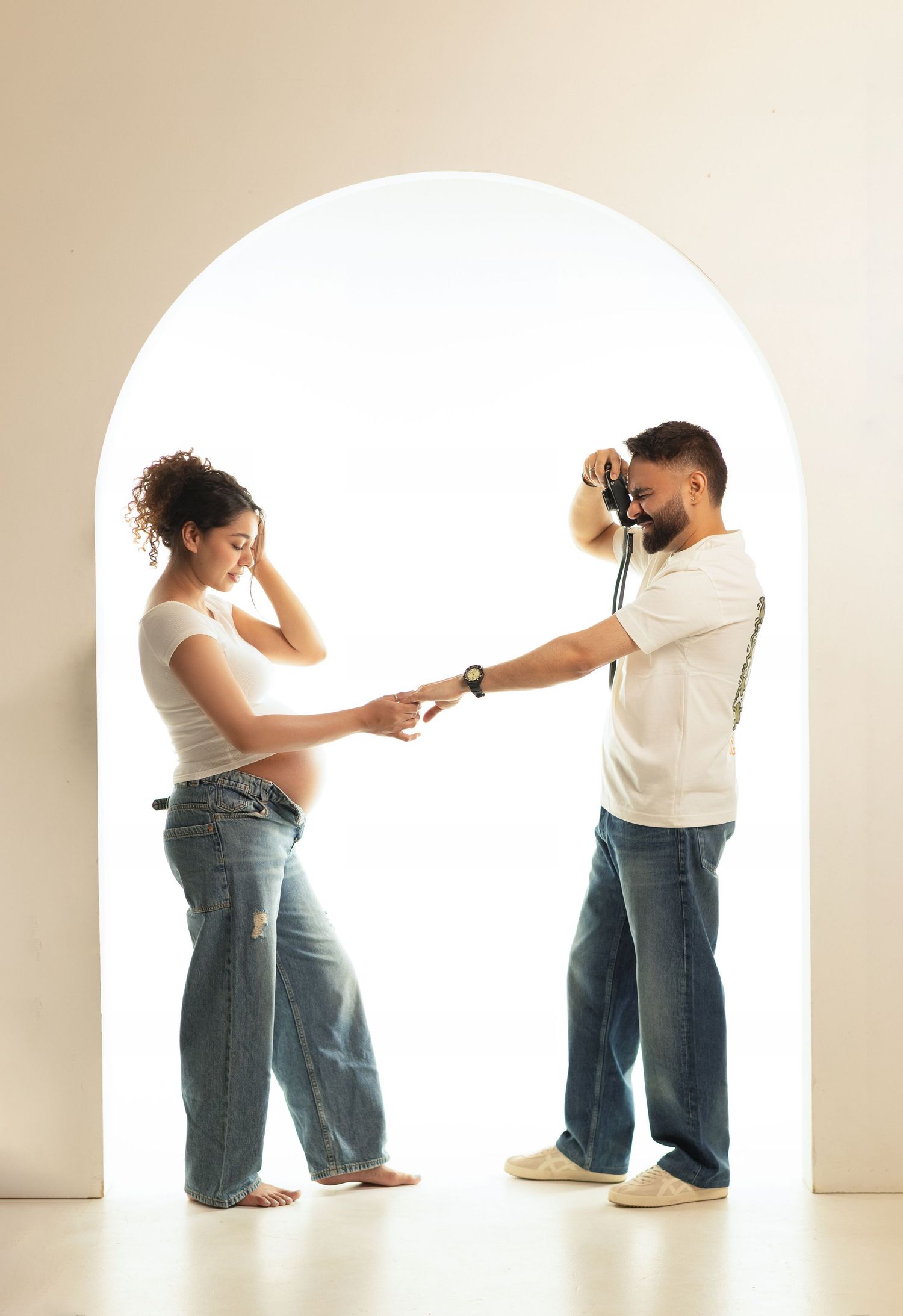 Pregnant woman and man holding hands in white arch doorway, both wearing jeans and white tops.