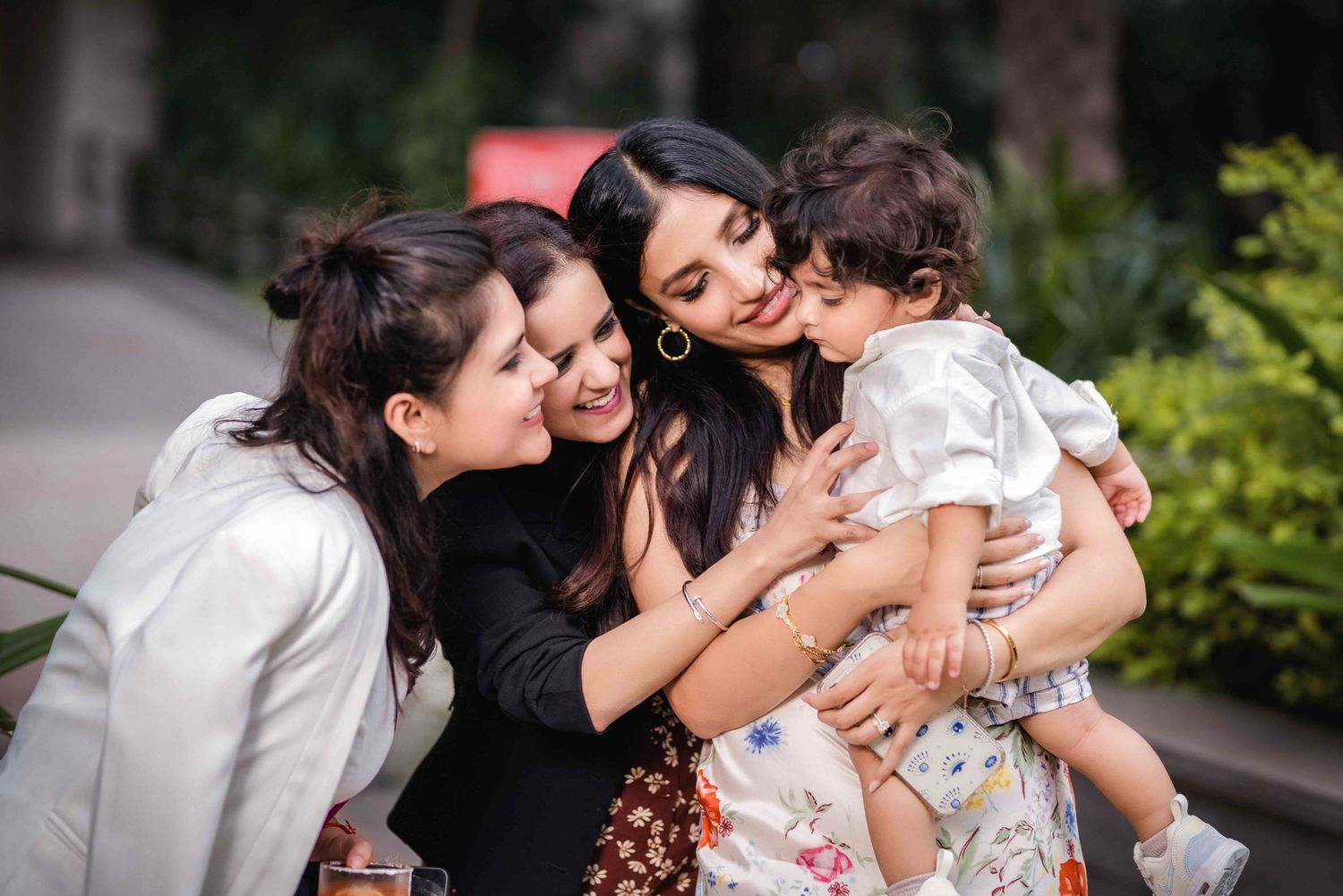 Three women smiling and kissing a cute toddler outdoors in a lush green setting.