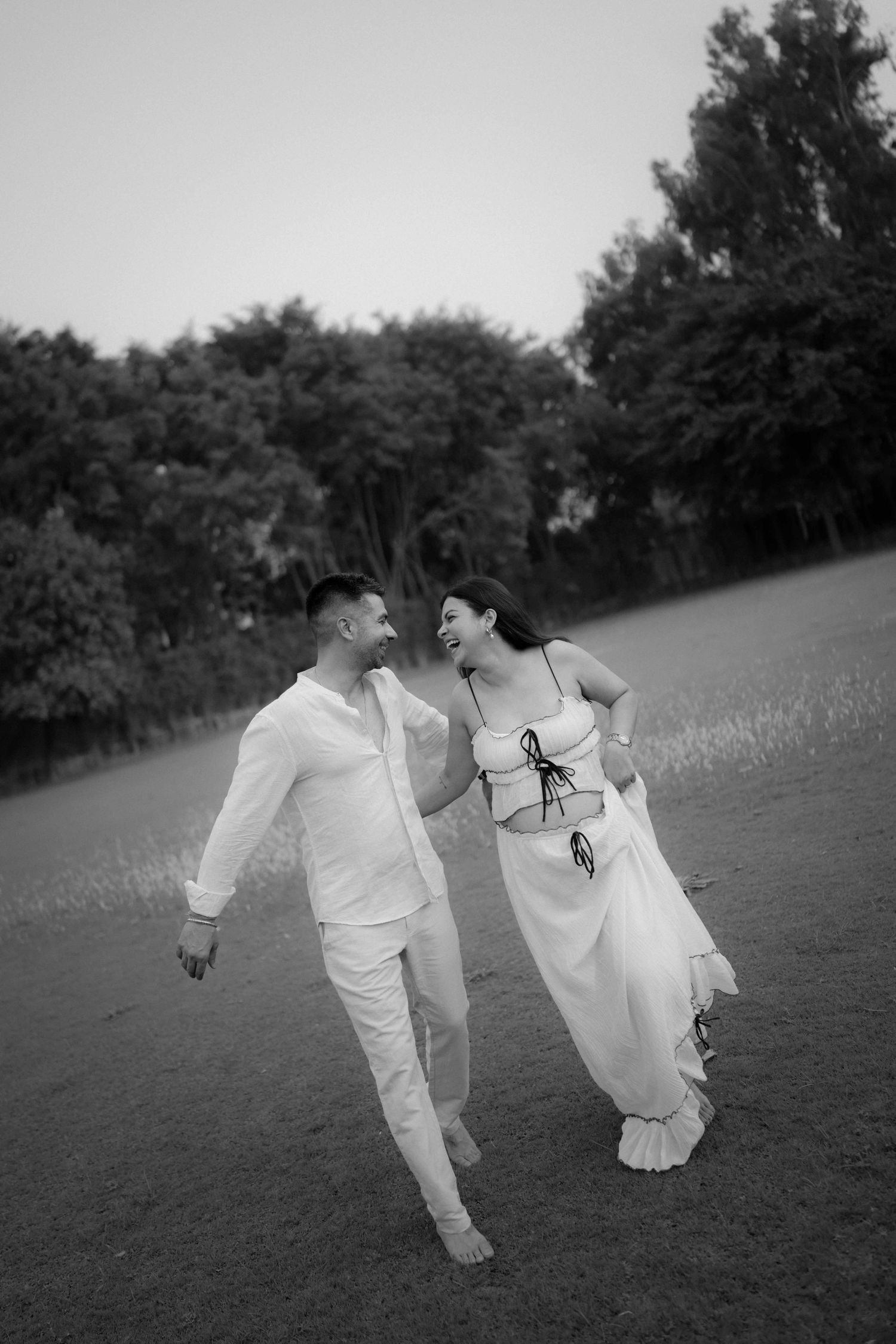 Black and white photo of a joyful couple in white outfits walking hand in hand through a lush green field.