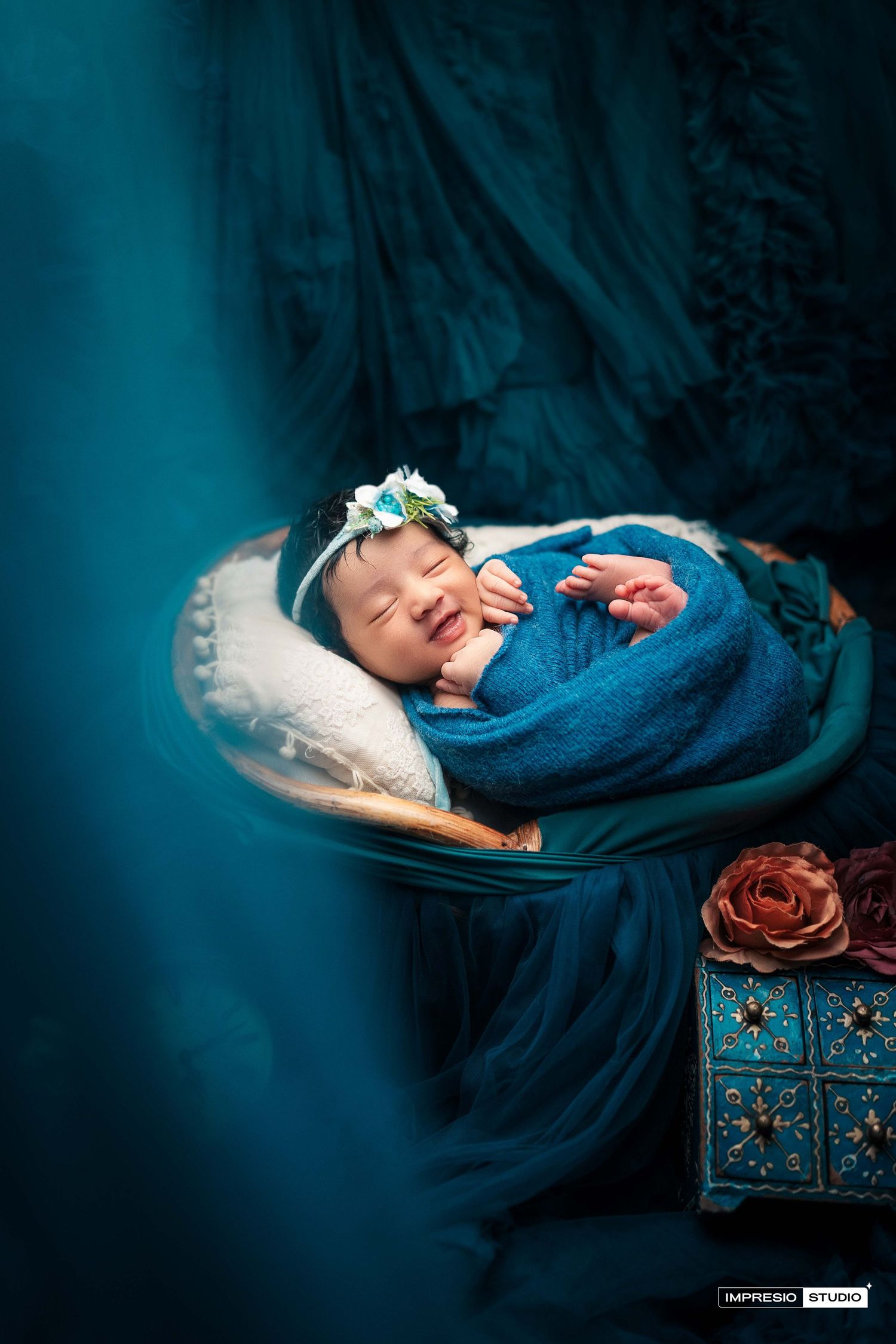 Smiling newborn baby wrapped in blue swaddle with floral headband in a teal-themed studio setting.