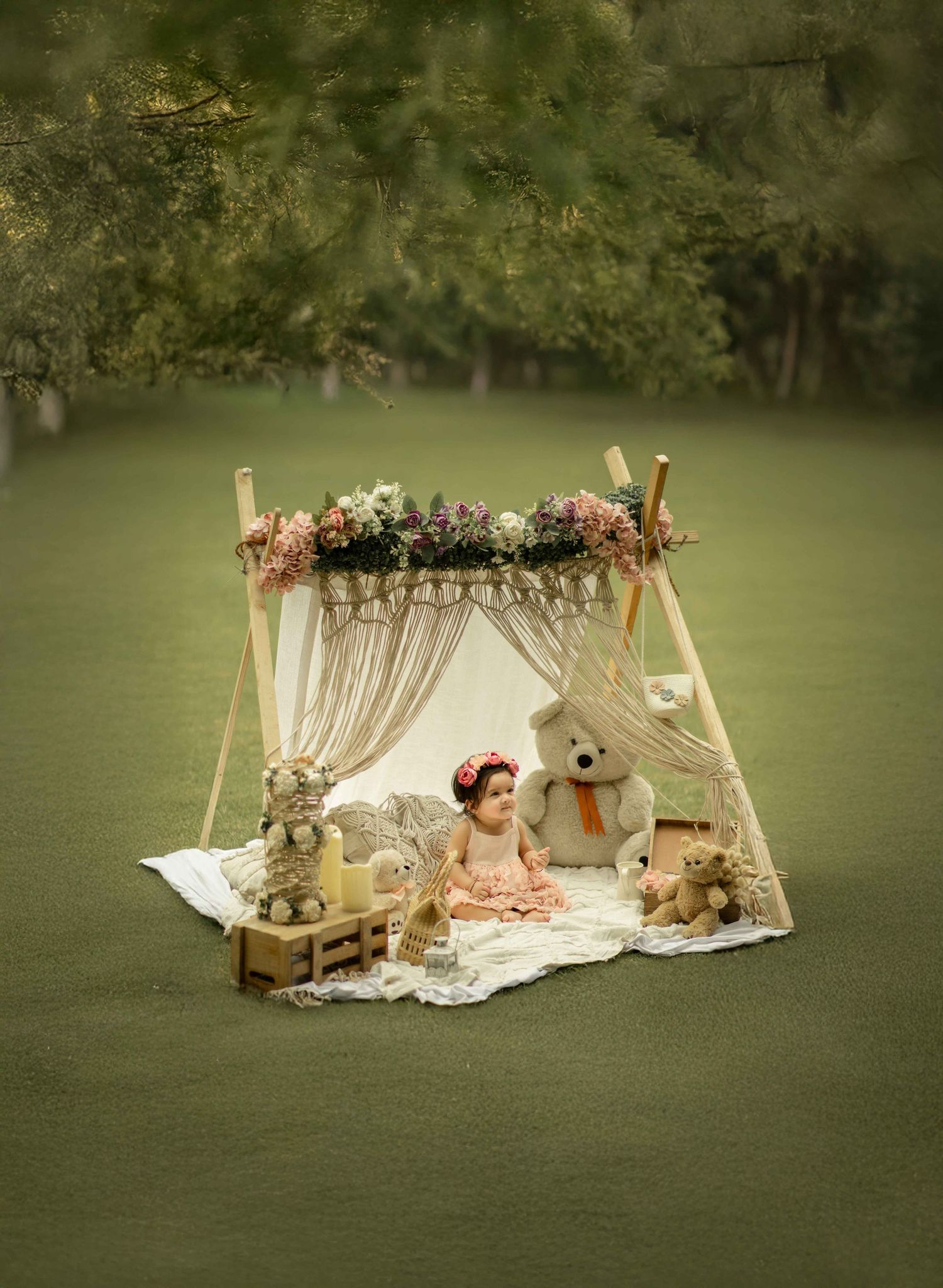 A baby girl sits inside a floral-topped boho tent with teddy bears on a white blanket in a lush green outdoor setting.