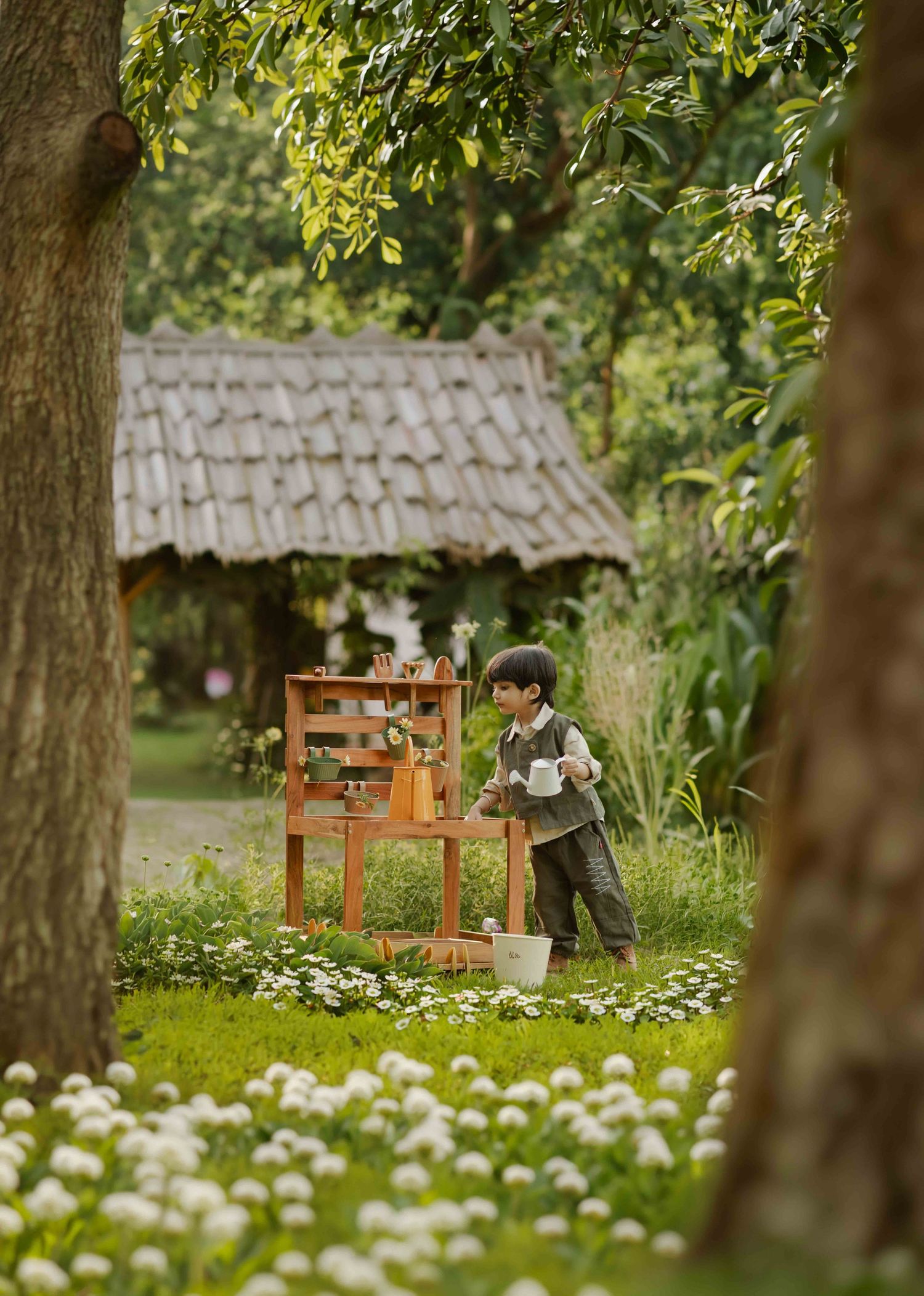 Young boy playing with wooden toy stand in lush garden with white flowers and rustic tiled roof structure.