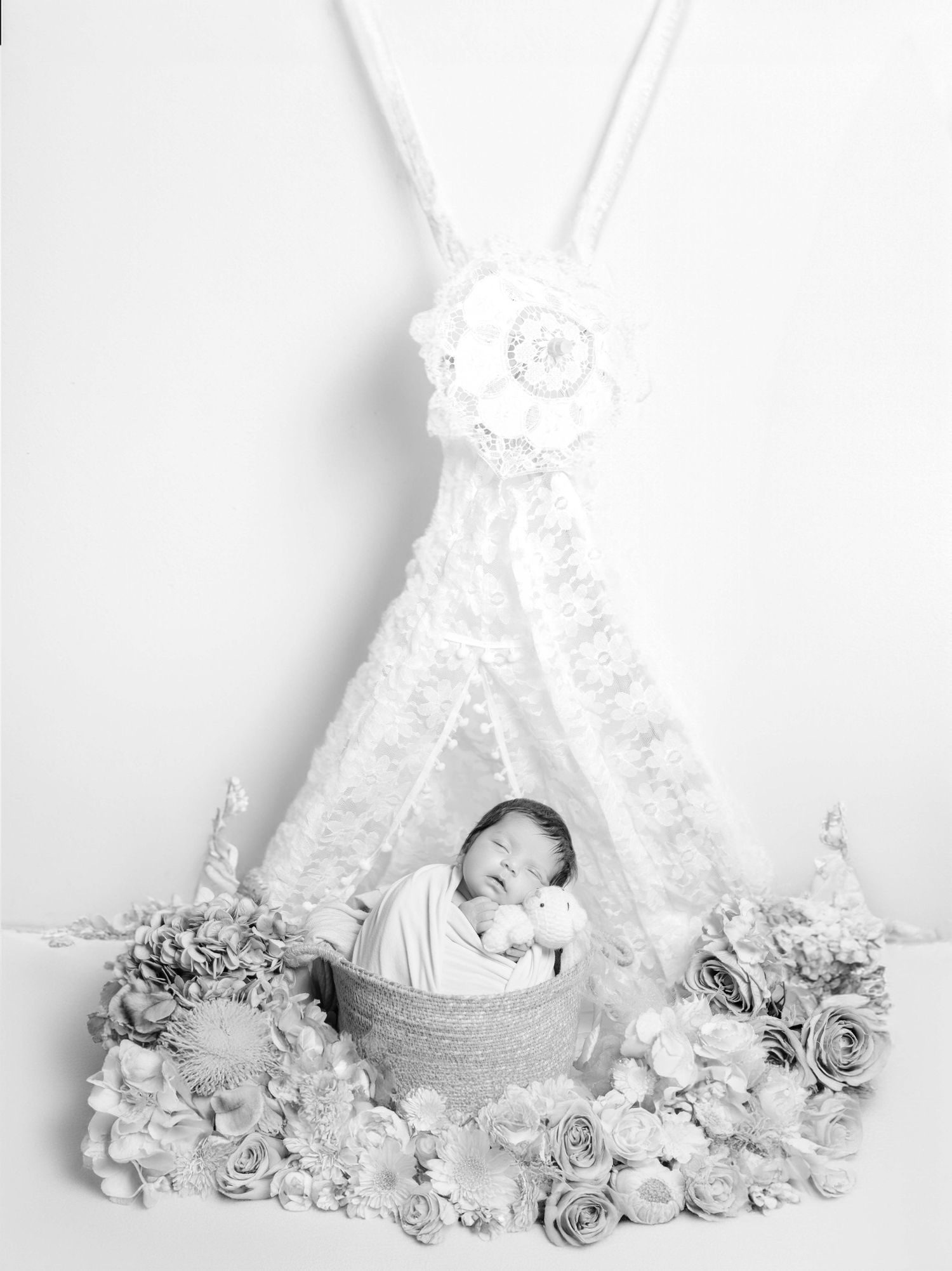 Black and white newborn photo of sleeping baby in basket surrounded by flowers beneath lace teepee.