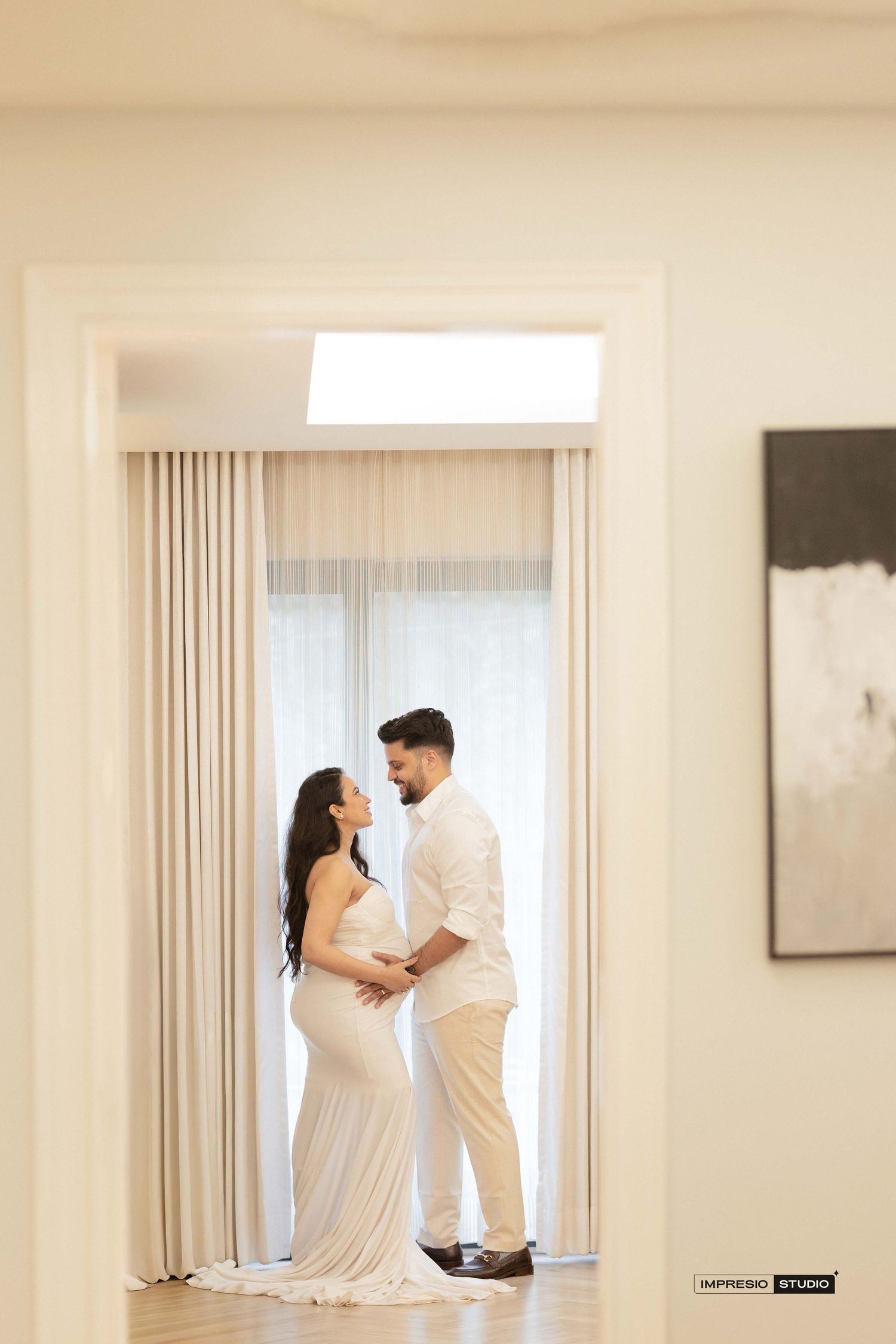 Couple in white outfits share a tender moment in an elegant, light-filled room during a maternity photoshoot.