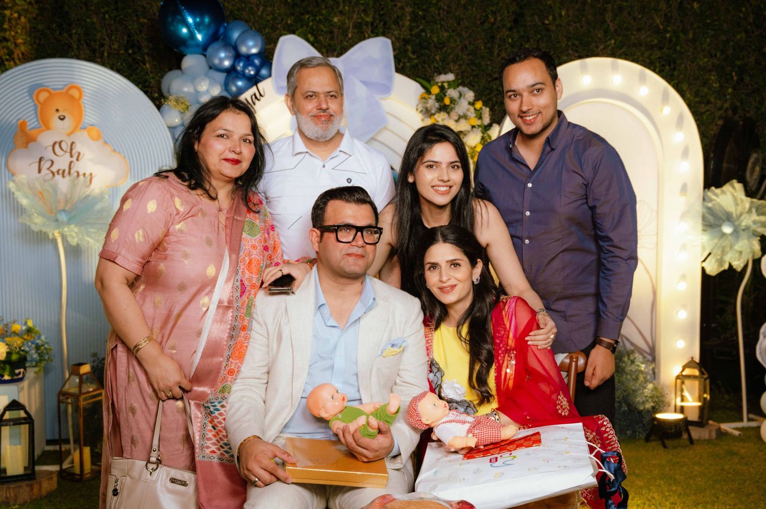 Group of six people posing together at a baby shower celebration with teddy bear backdrop and balloon decorations.