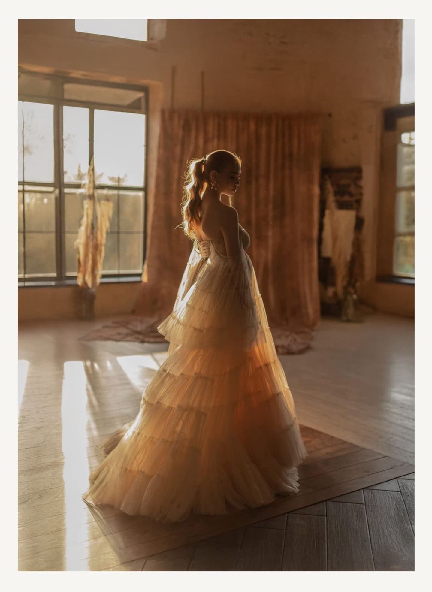 A silhouetted figure stands in a sunlit room wearing a flowing tiered wedding gown with soft tulle layers.