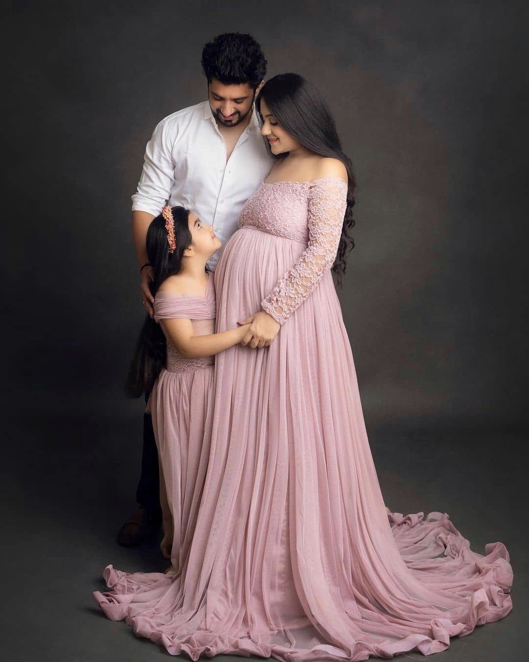 A couple poses lovingly against a dark background in a flowy pink maternity gown for a professional photoshoot.