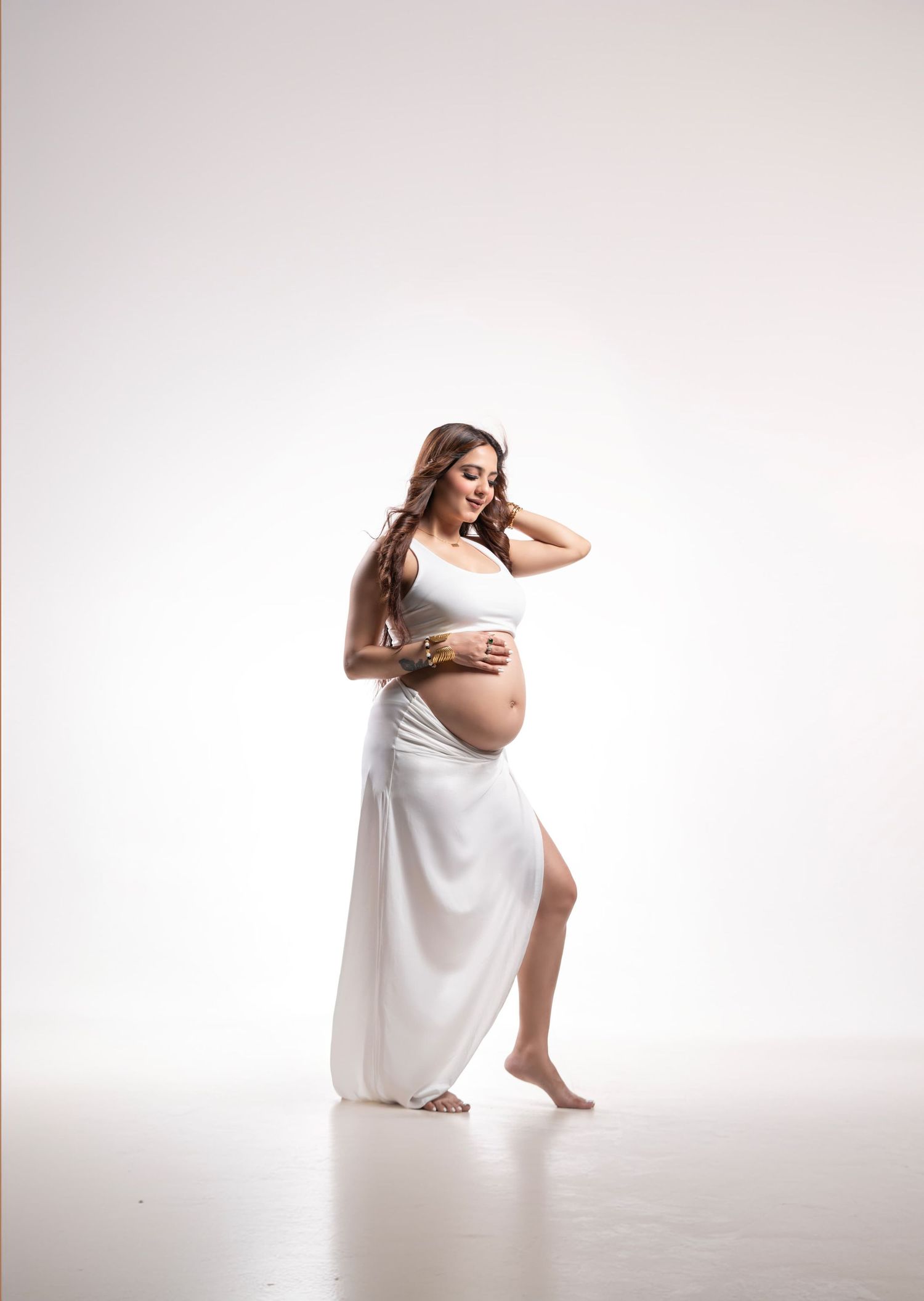Minimalist maternity portrait in white dress against light background with graceful pose.