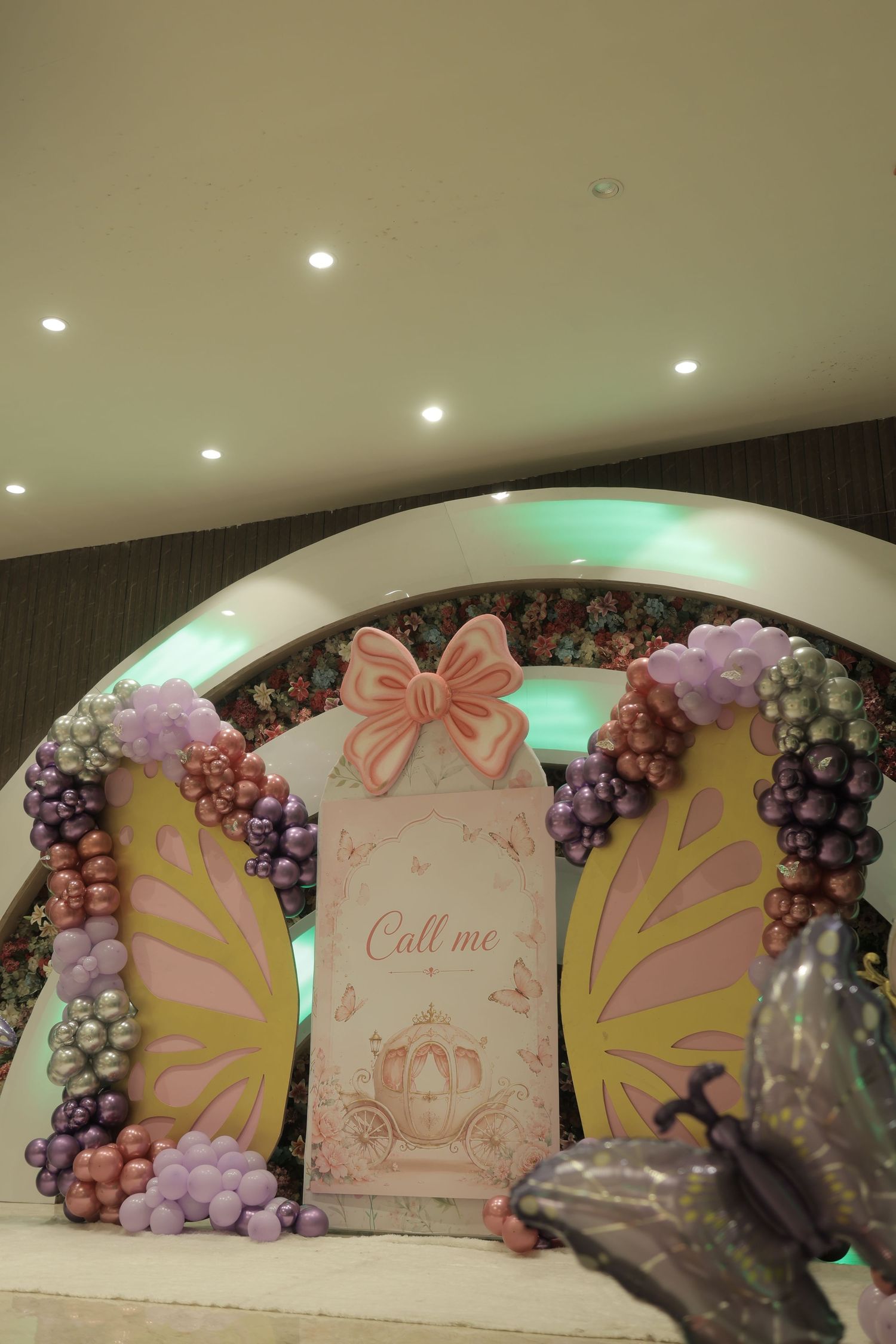 Butterfly-themed party backdrop with pink and gold wings, purple and mauve balloon garlands, a bow-topped carriage sign and a floral arch.