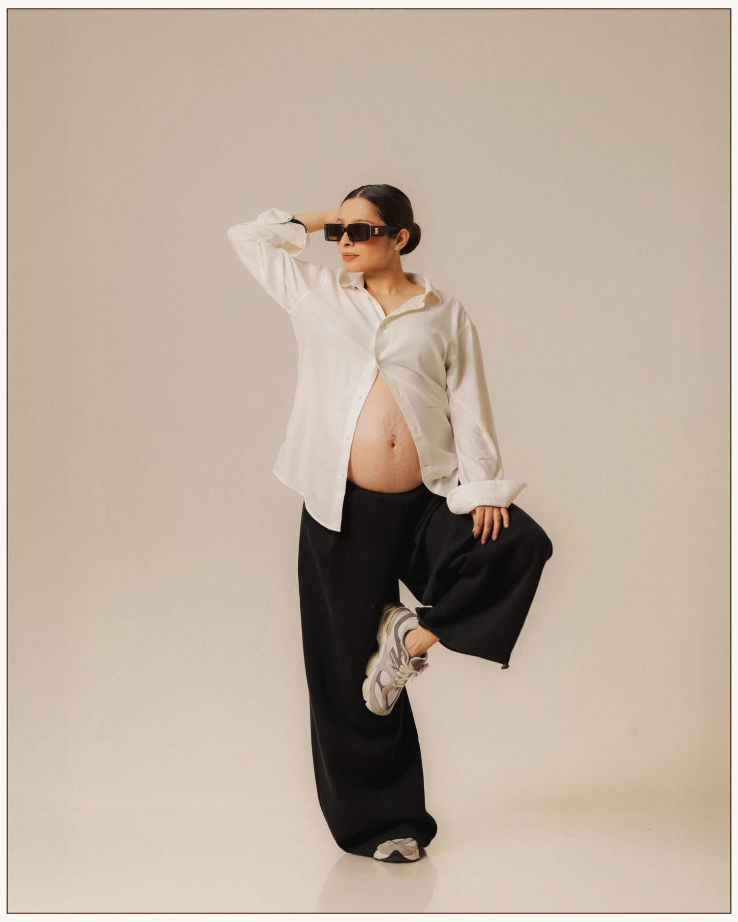 Pregnant woman in white shirt and black wide-leg pants poses confidently wearing sunglasses, showing baby bump.