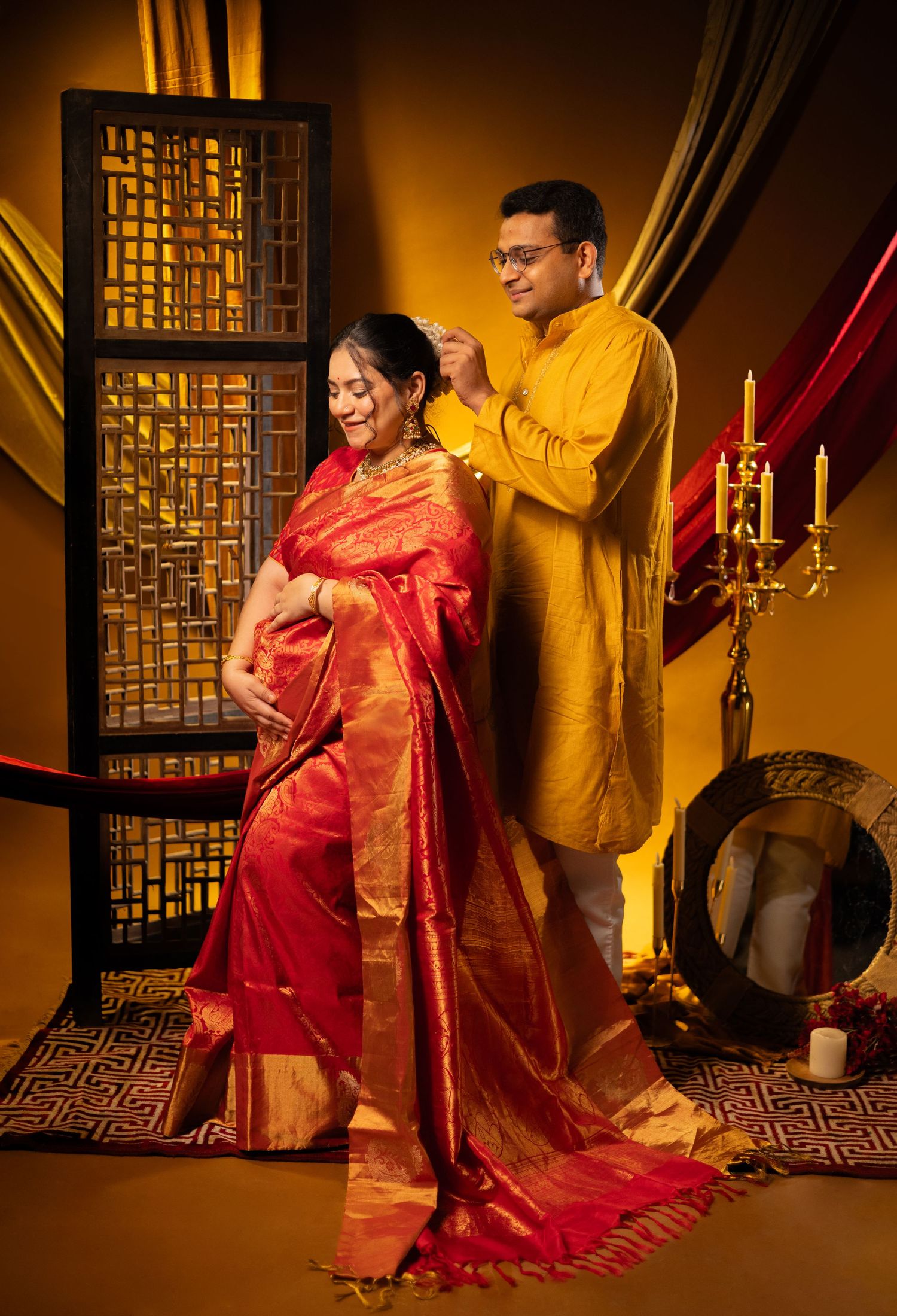 Traditional Indian couple in festive attire, wearing yellow kurta and red silk saree, pose against warm golden backdrop.