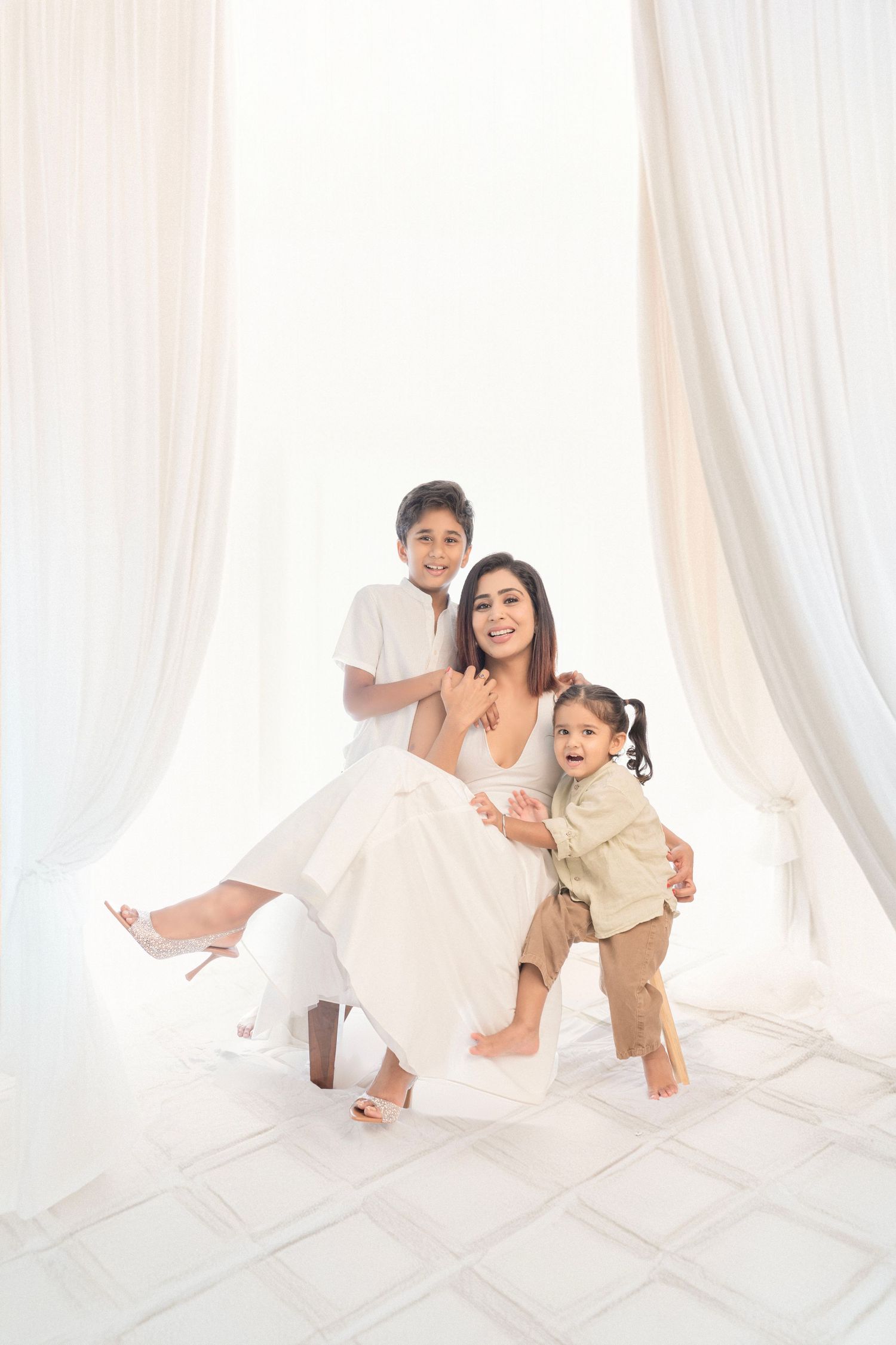 A smiling woman in white dress poses with two young children in a bright white room with flowing curtains.