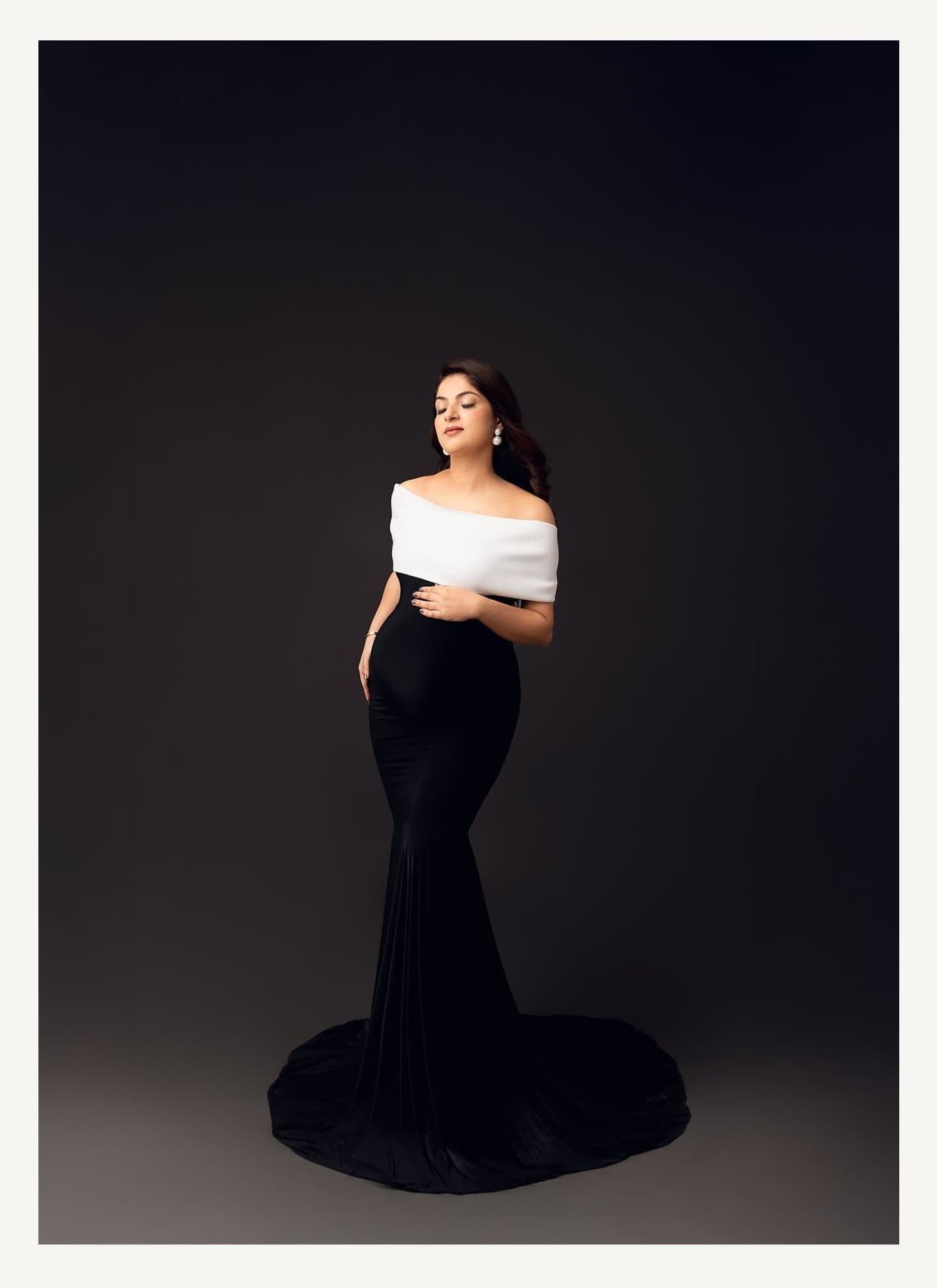 Dramatic maternity portrait in a black mermaid gown with white off-shoulder detail against dark background.