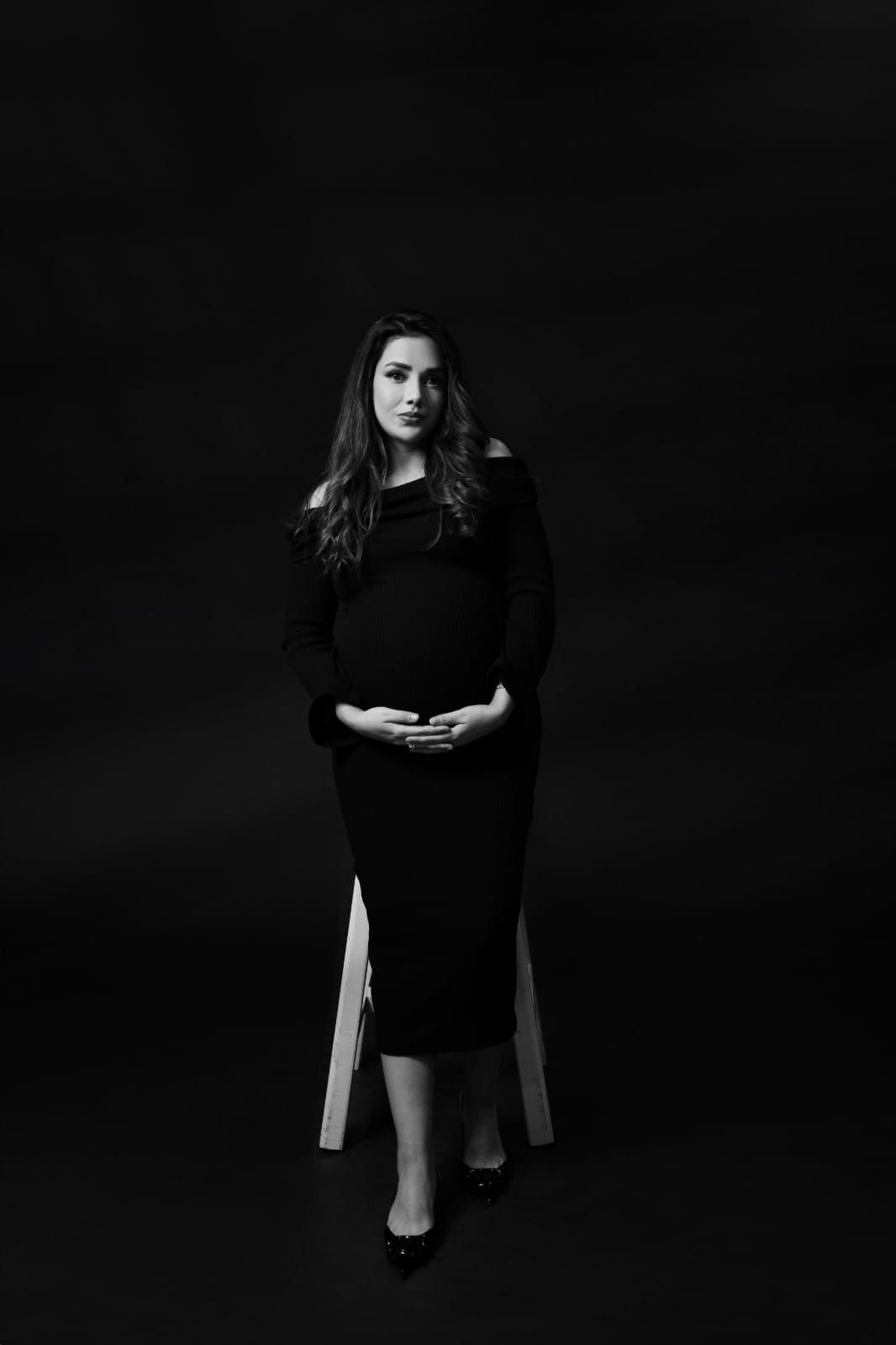 Dramatic black and white maternity portrait featuring black dress against dark background.
