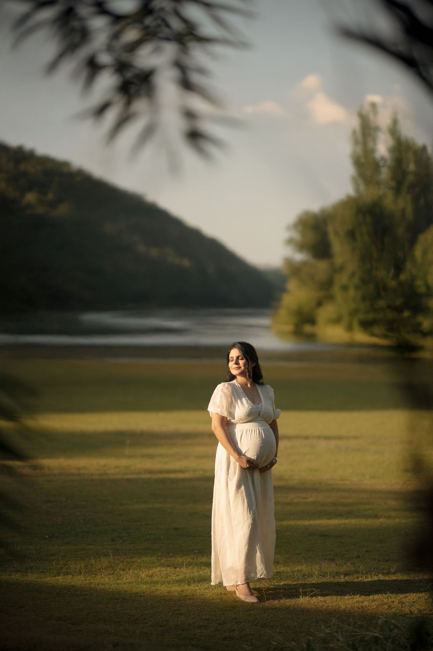 Outdoor maternity photoshoot flowing white dress stands by a serene lake at sunset, cradling their belly in a peaceful natural setting.