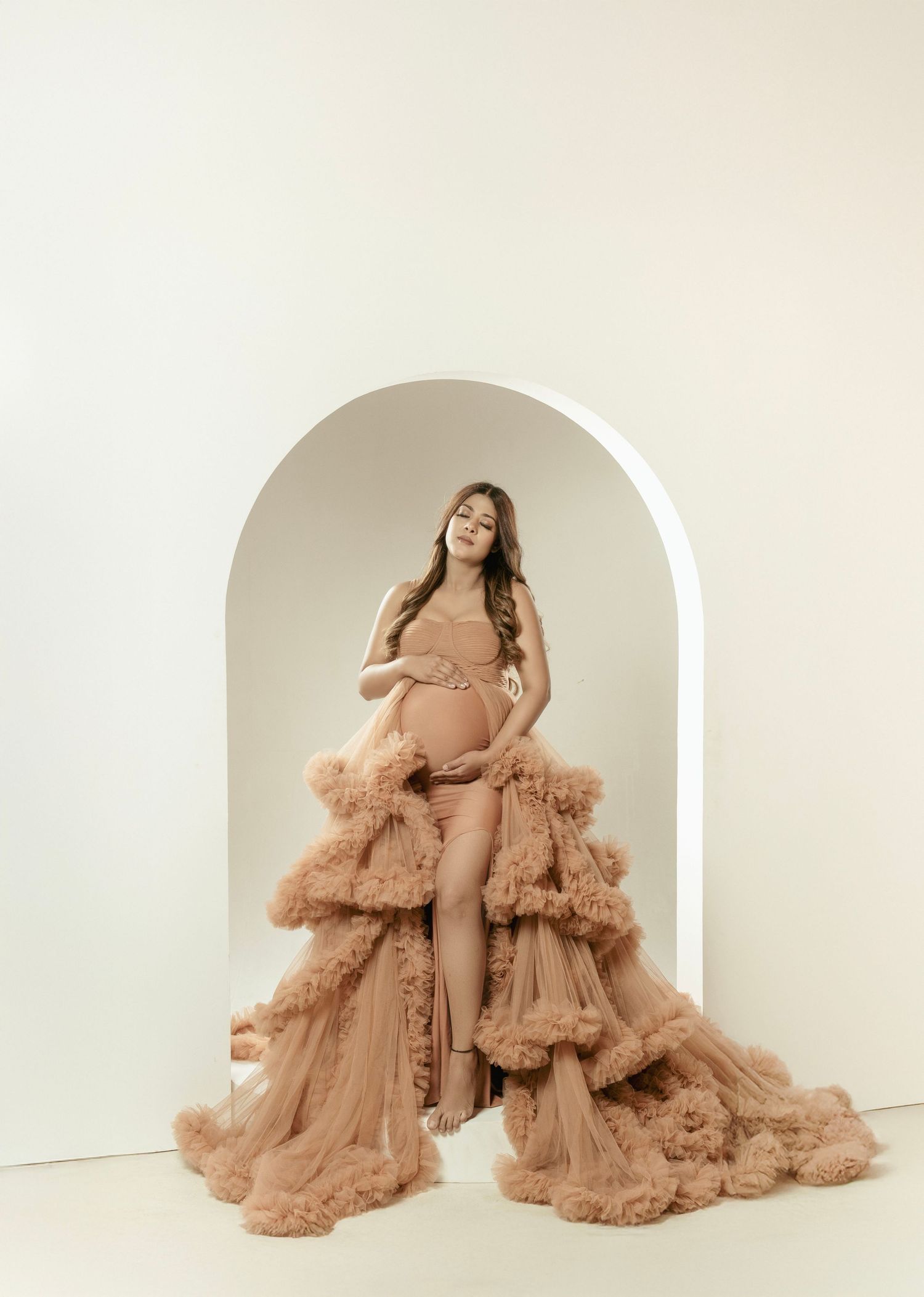 Ethereal maternity photoshoot featuring flowing dusty rose tulle gown in minimalist arch setting.