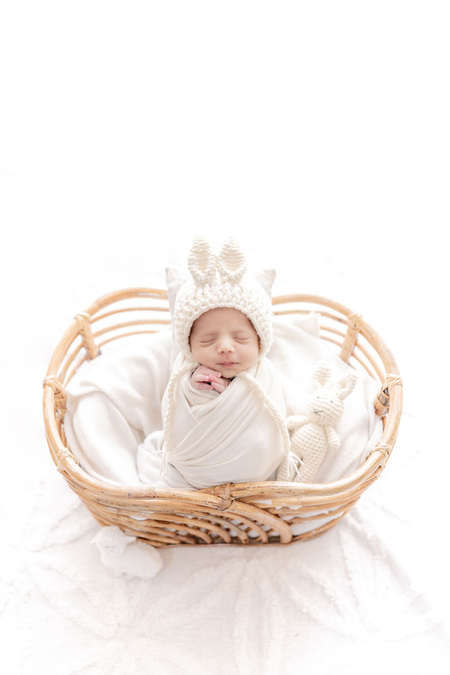 A sleeping newborn baby wearing a knitted crown hat lies peacefully wrapped in white cloth in a wicker basket.