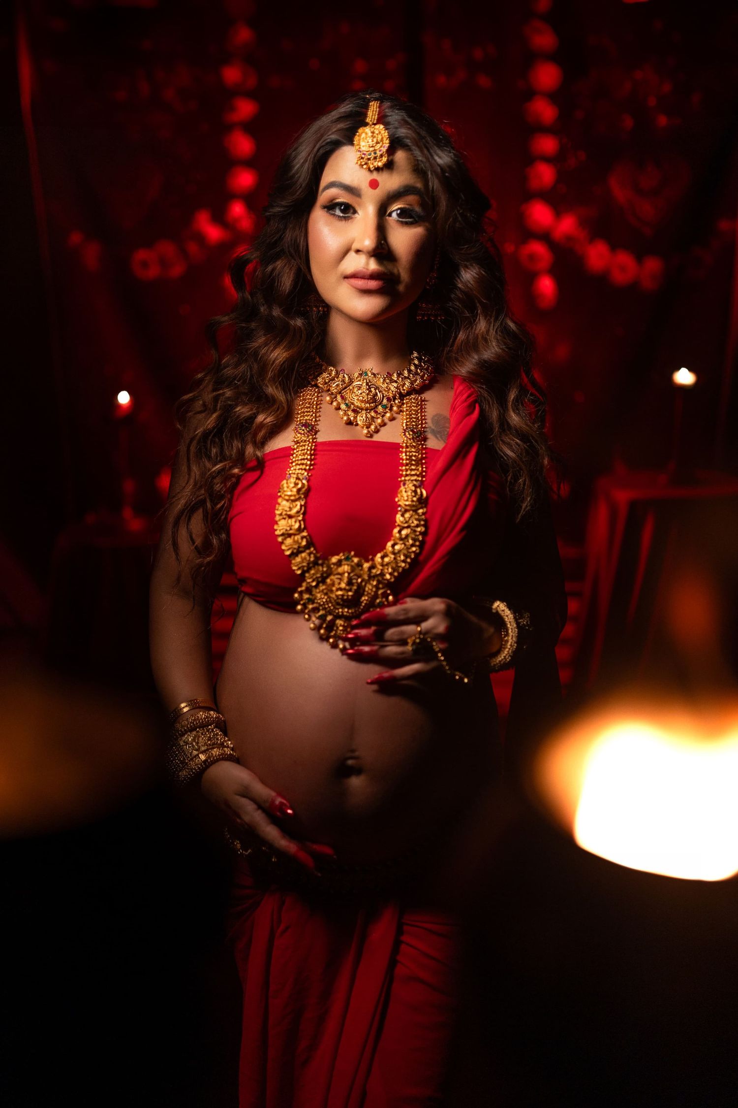 A figure in a red dress and gold jewelry poses in a dramatic moody setting with red lighting and shadows for maternity photography