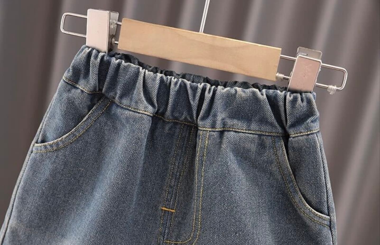 Three pairs of light blue and medium wash denim jeans hanging on wooden hangers against a gray background.