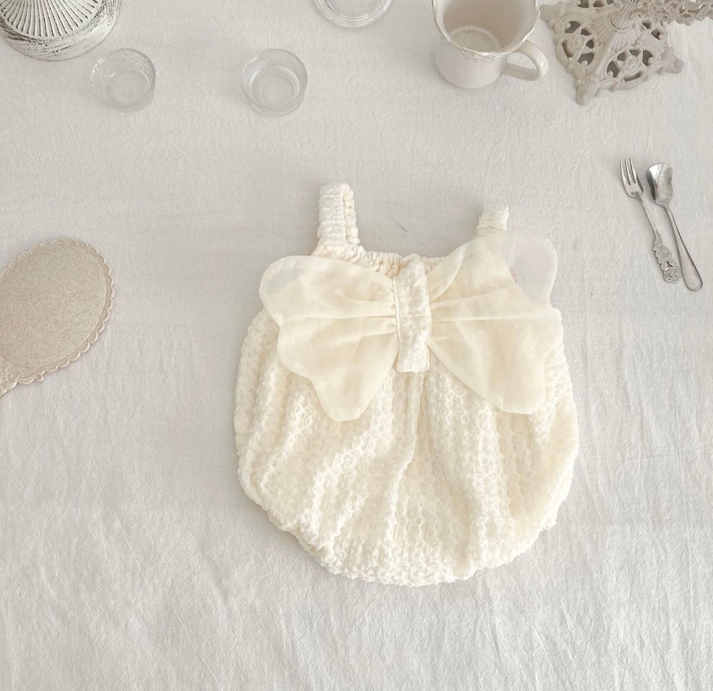 Delicate cream-colored baby romper with eyelet lace details and a large bow accent laid flat on white linens.