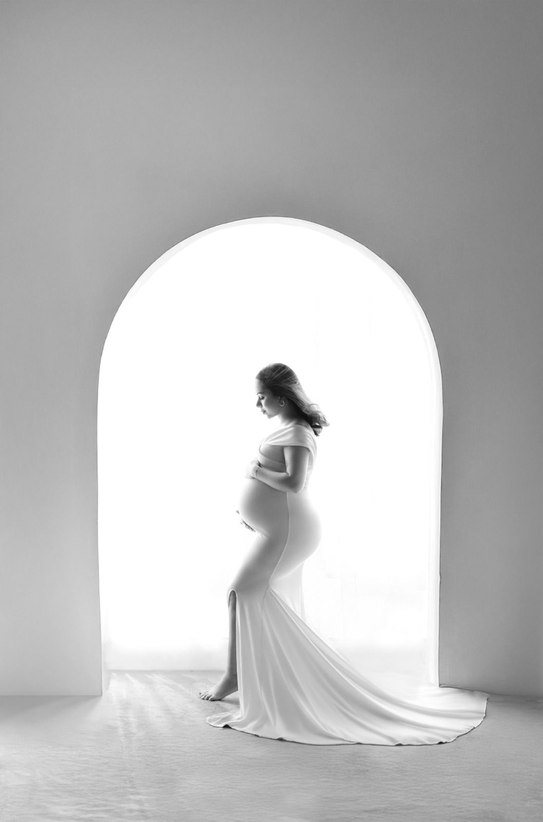 Black and white maternity photo of pregnant woman in elegant white gown posing in front of arched doorway.