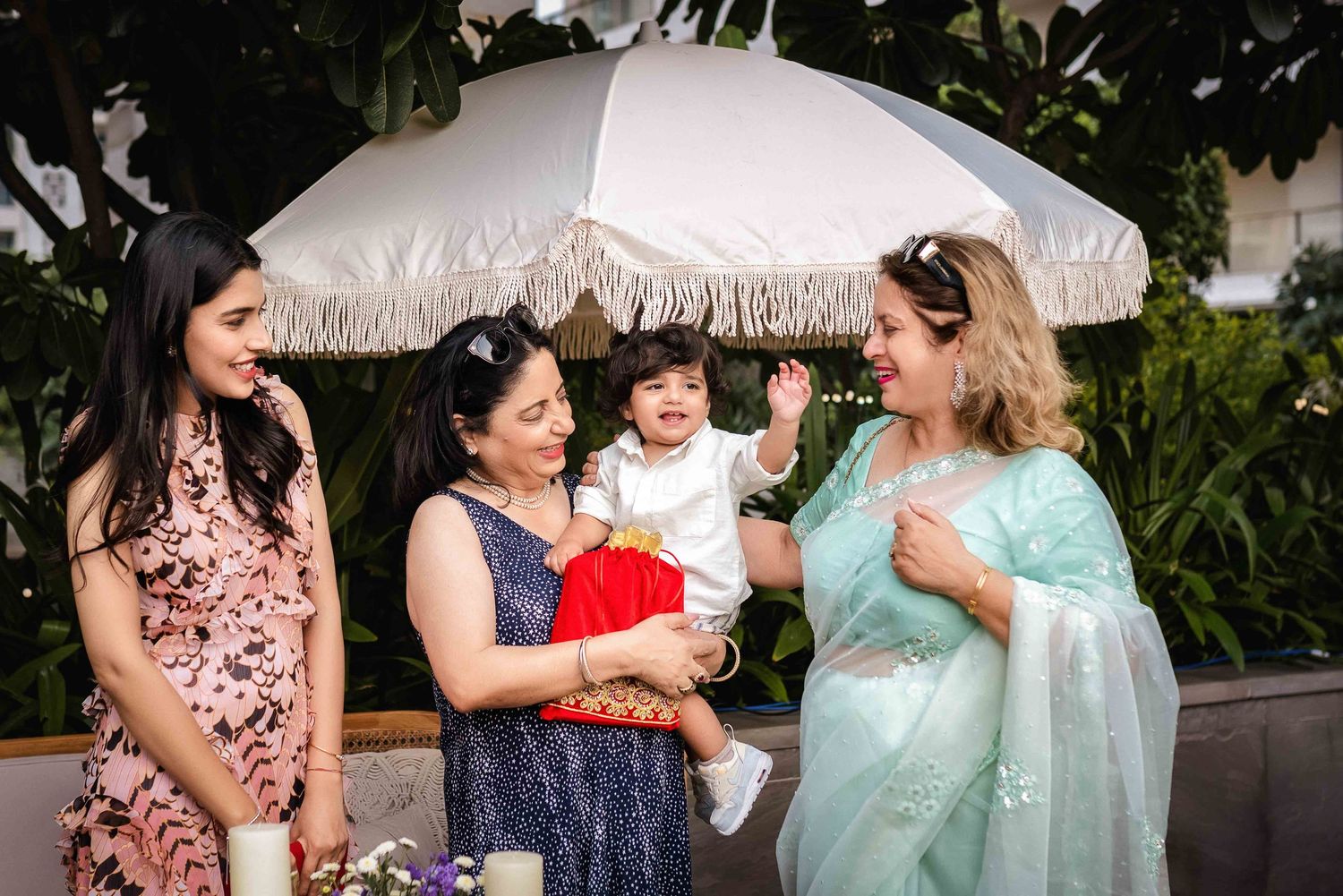 Three women and a baby celebrate outdoors under a white fringed umbrella at an Indian cultural event.