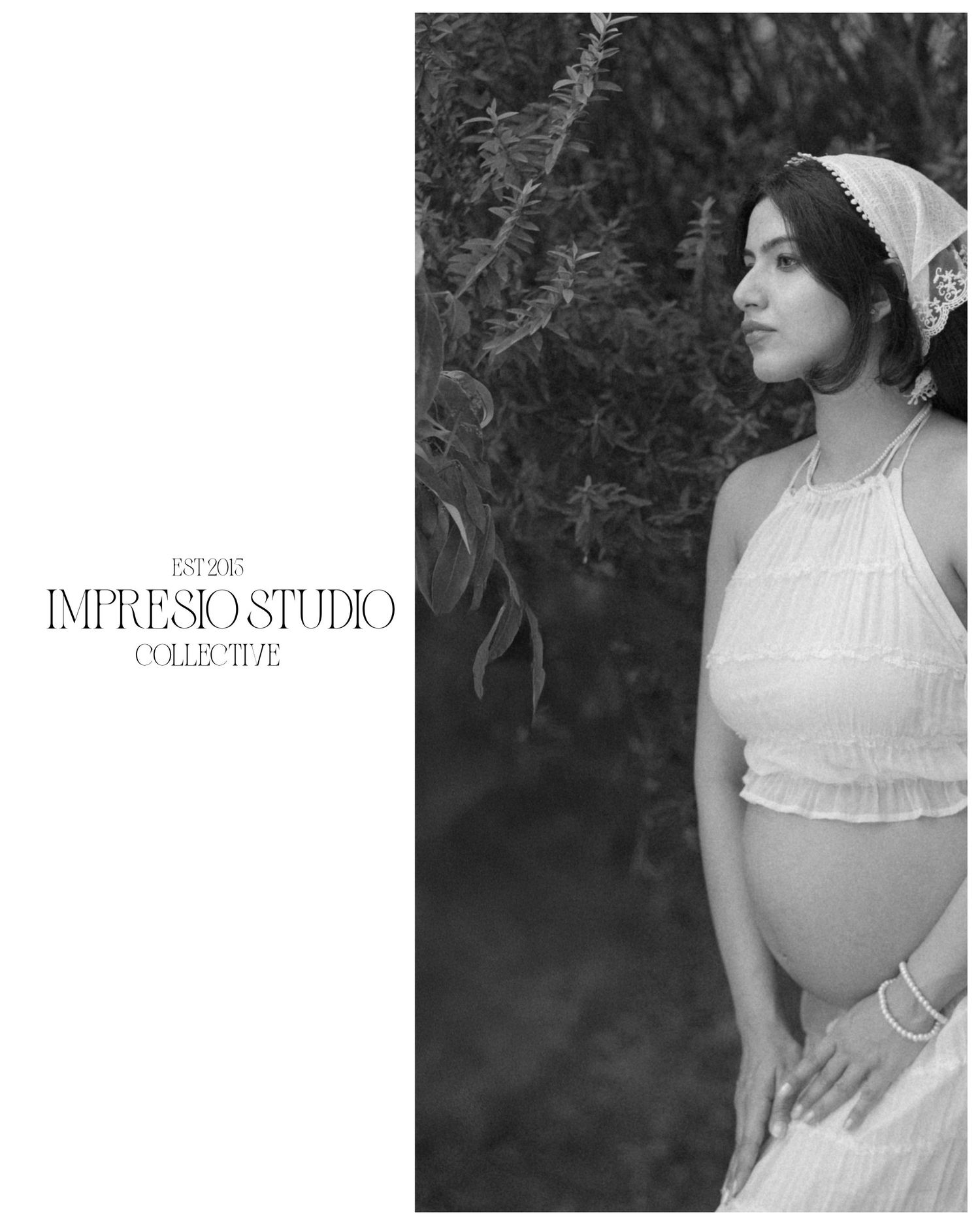 Black and white maternity photo of a woman in a white halter dress and headband, posed outdoors by lush greenery.