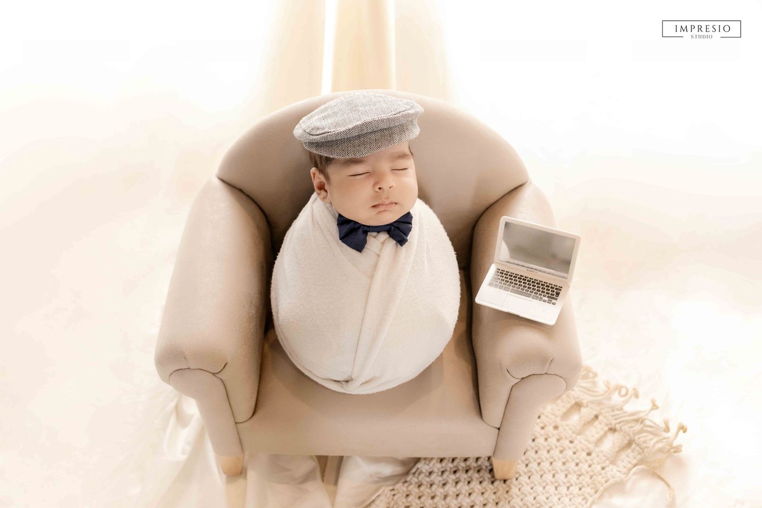 Sleeping newborn baby wrapped in white, wearing a flat cap and bow tie, posed in a miniature armchair with a tiny laptop.
