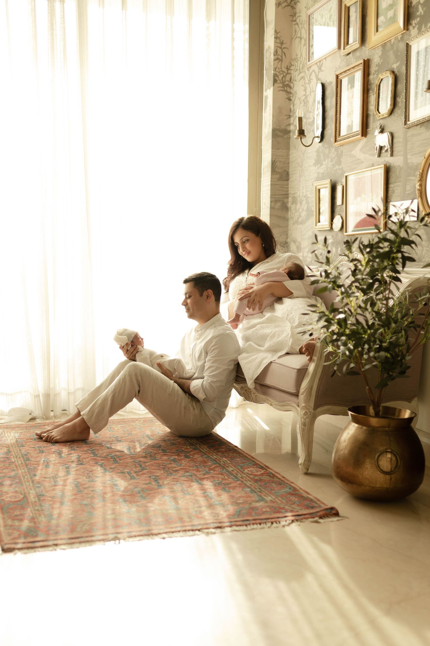 A couple sits peacefully in a sunlit room, the woman holding a newborn baby while the man sits on a patterned rug.