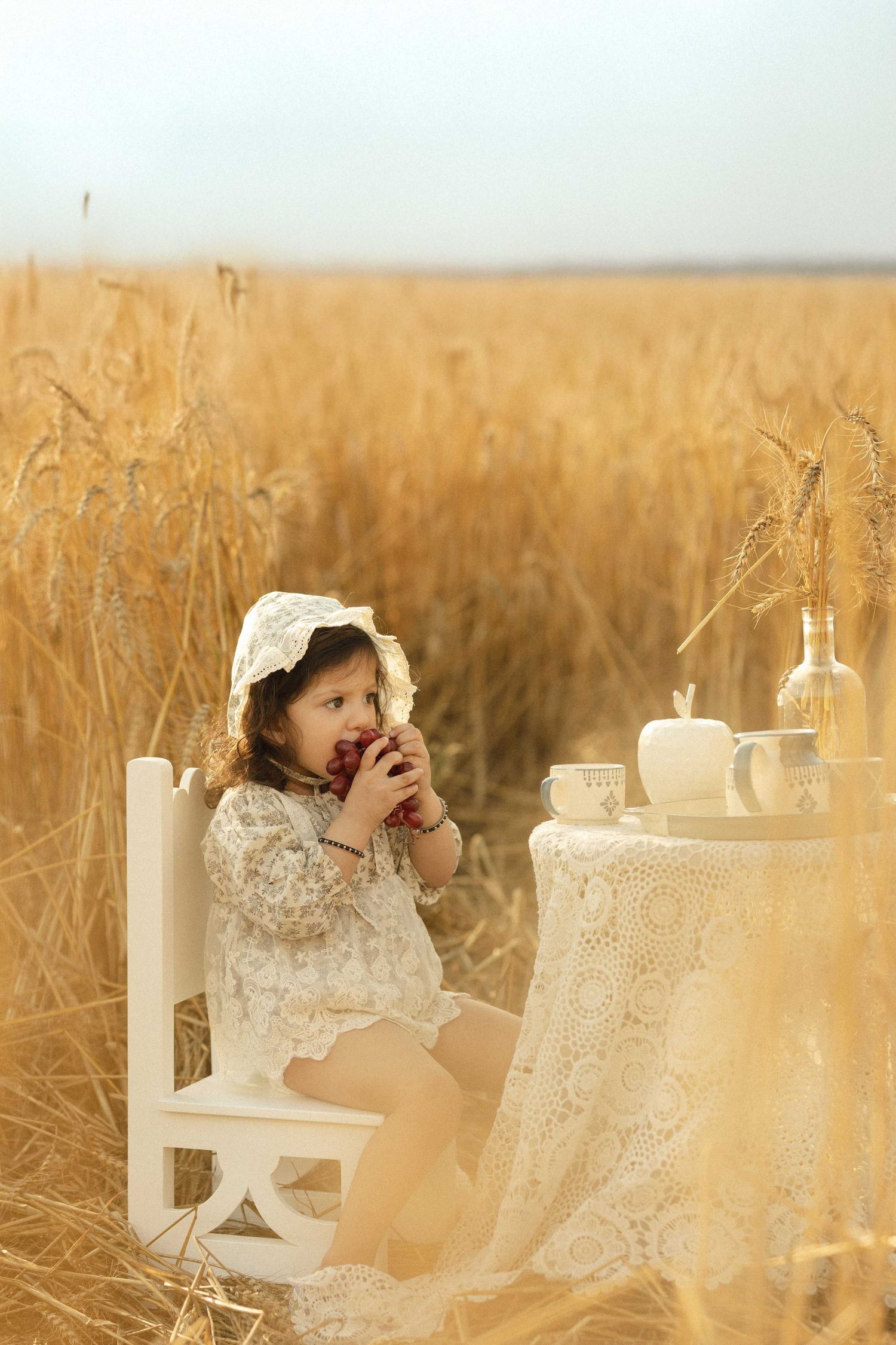 A little girl in a lace dress and bonnet sits in a wheat field eating an apple at a small white table.
