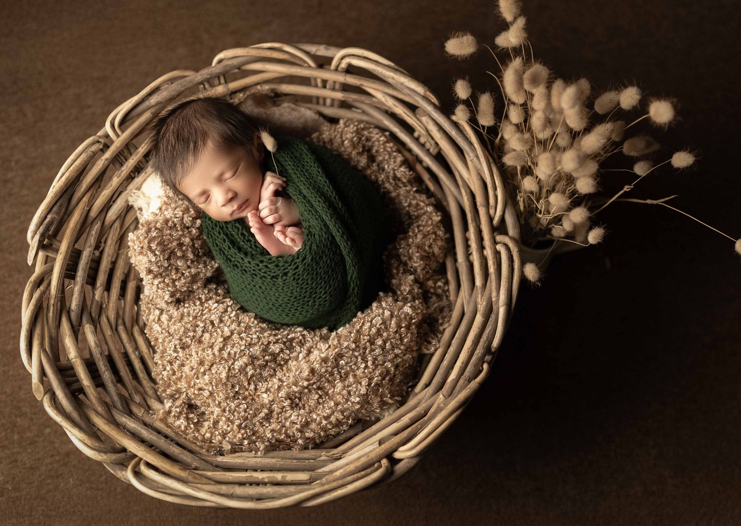 Sleeping newborn wrapped in green knit swaddle nestled in a wicker basket with decorative dried flowers nearby.