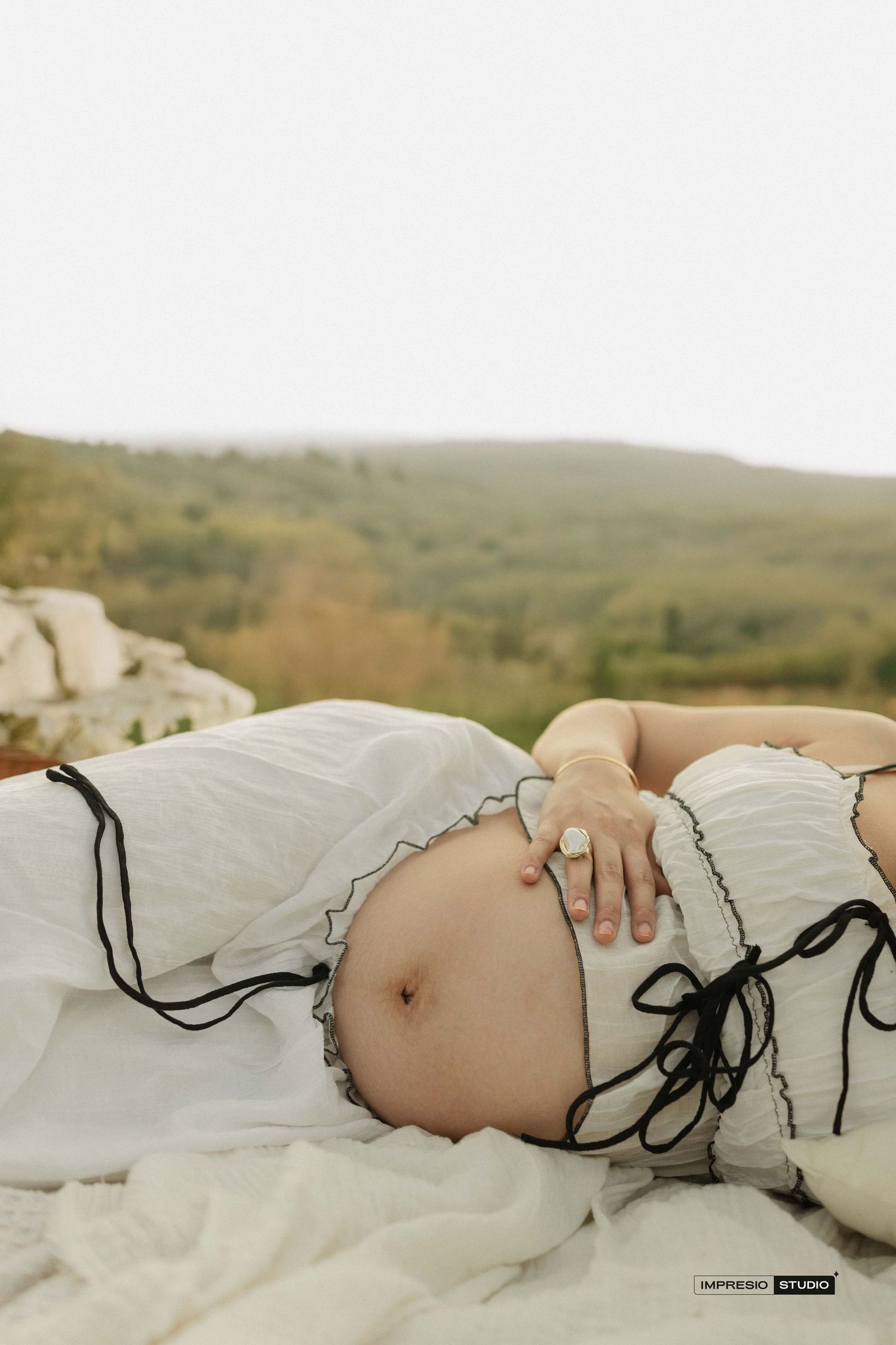Pregnant woman lying outdoors in white dress with black ties, showing baby bump against scenic green hillside backdrop.