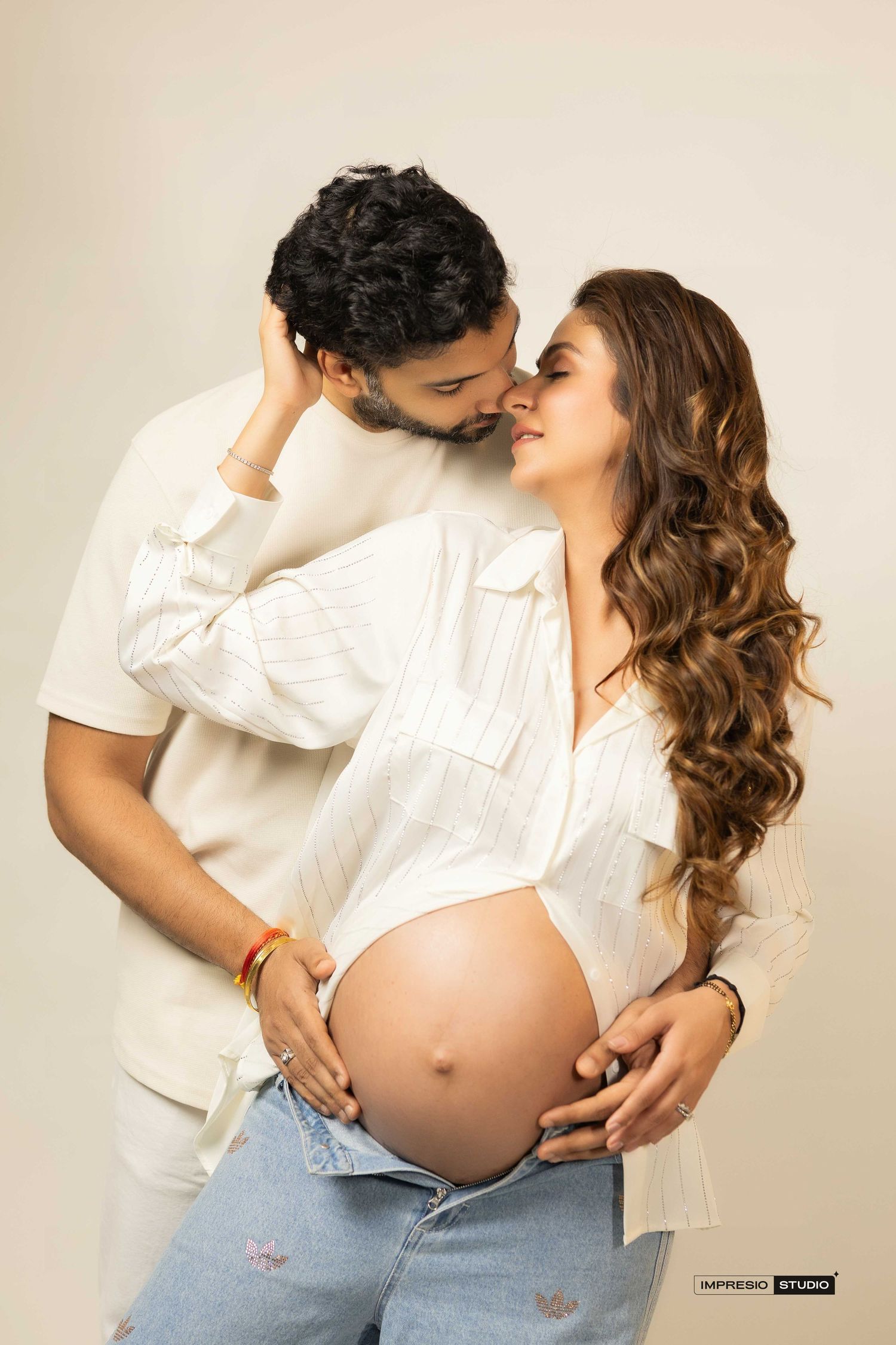 Couple in white shirts posing for maternity photoshoot, woman showing baby bump, studio photography.