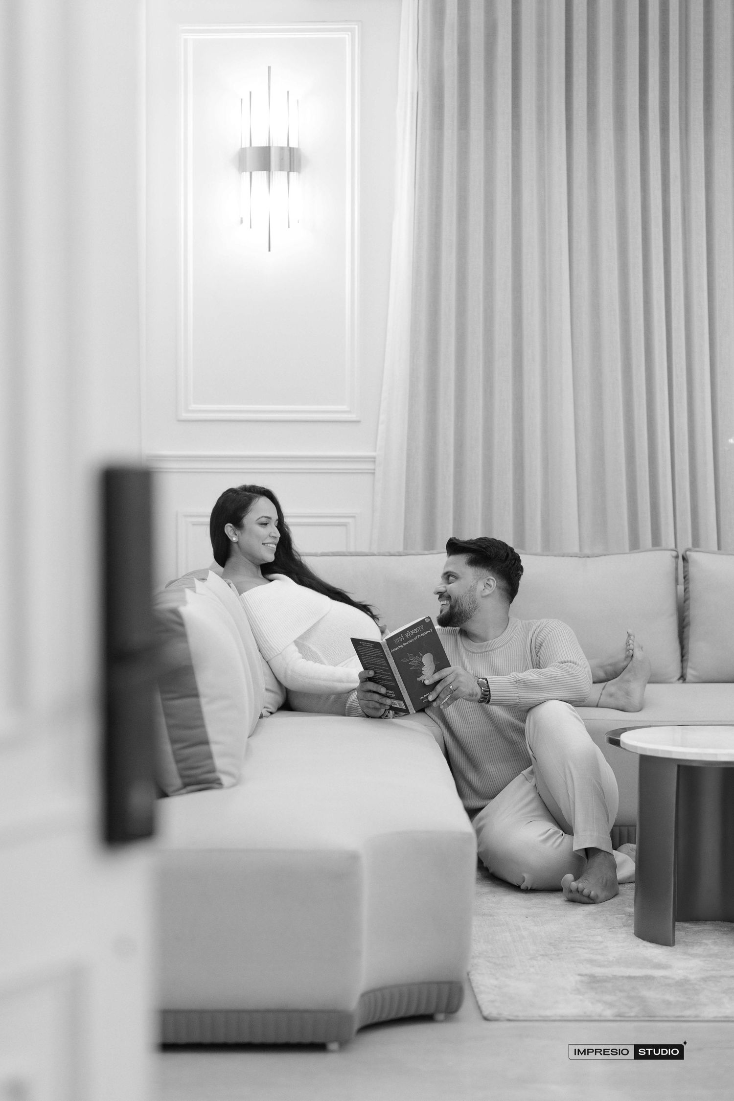 A couple relaxes on a sofa, sharing a book in a bright, elegant living room in this black and white photo.