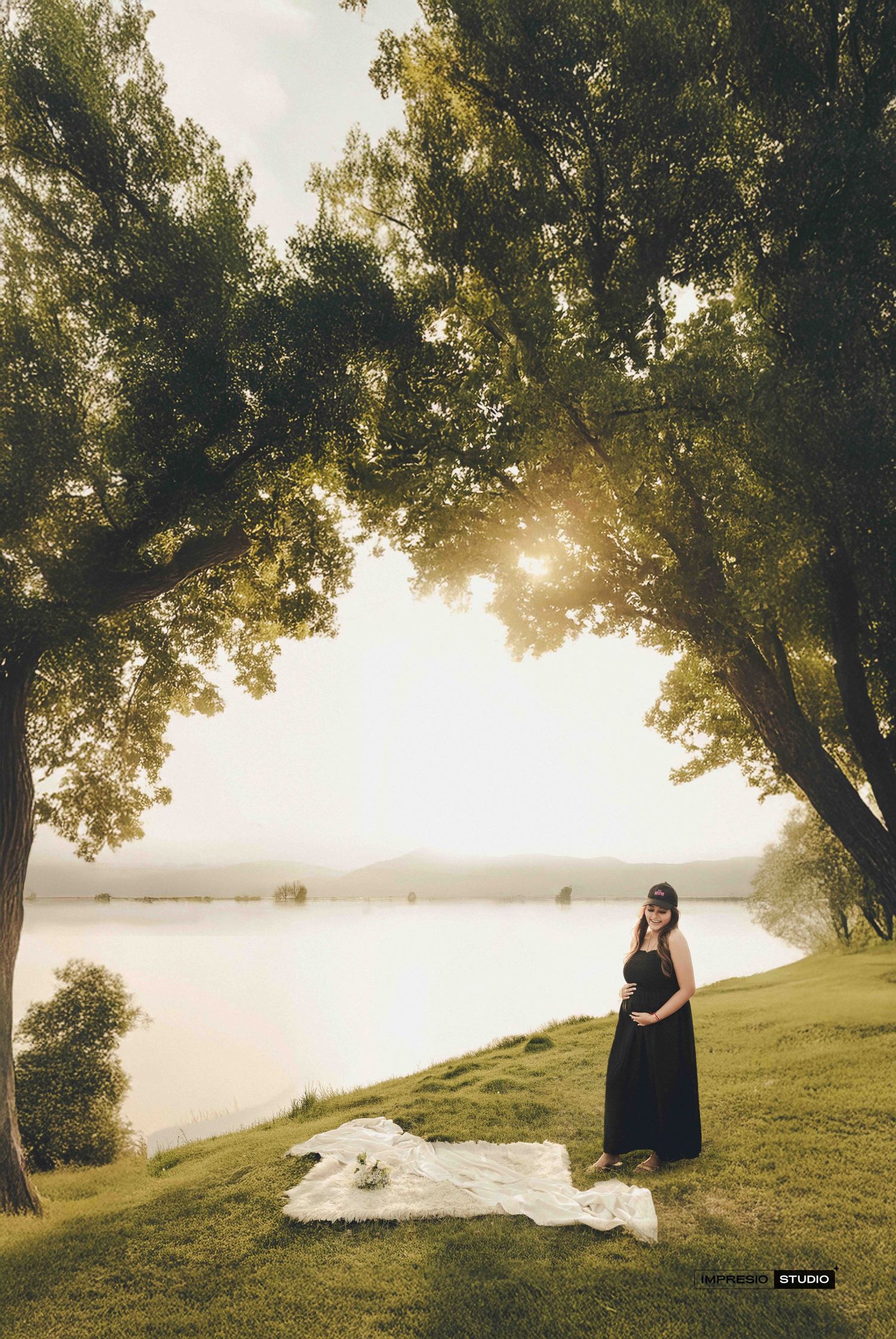 Pregnant woman in black dress standing by a serene lake under lush green trees during golden hour maternity photoshoot.