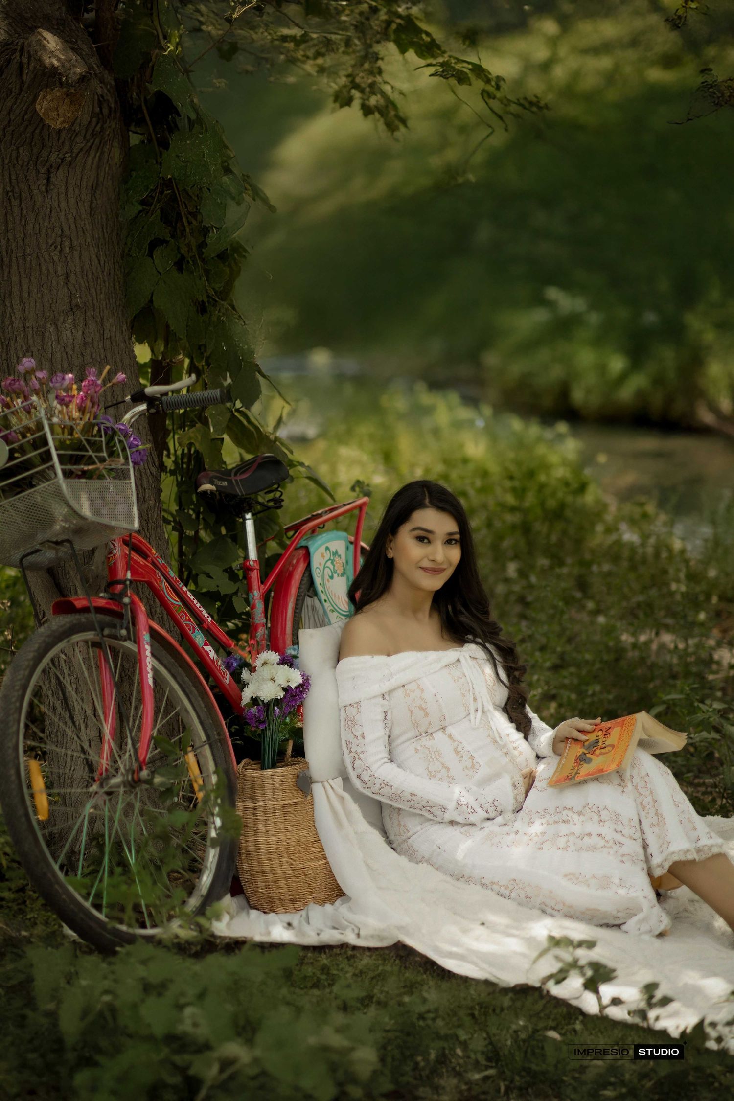 Pregnant woman in white lace dress sits under tree with red bicycle and flower basket in lush garden setting.