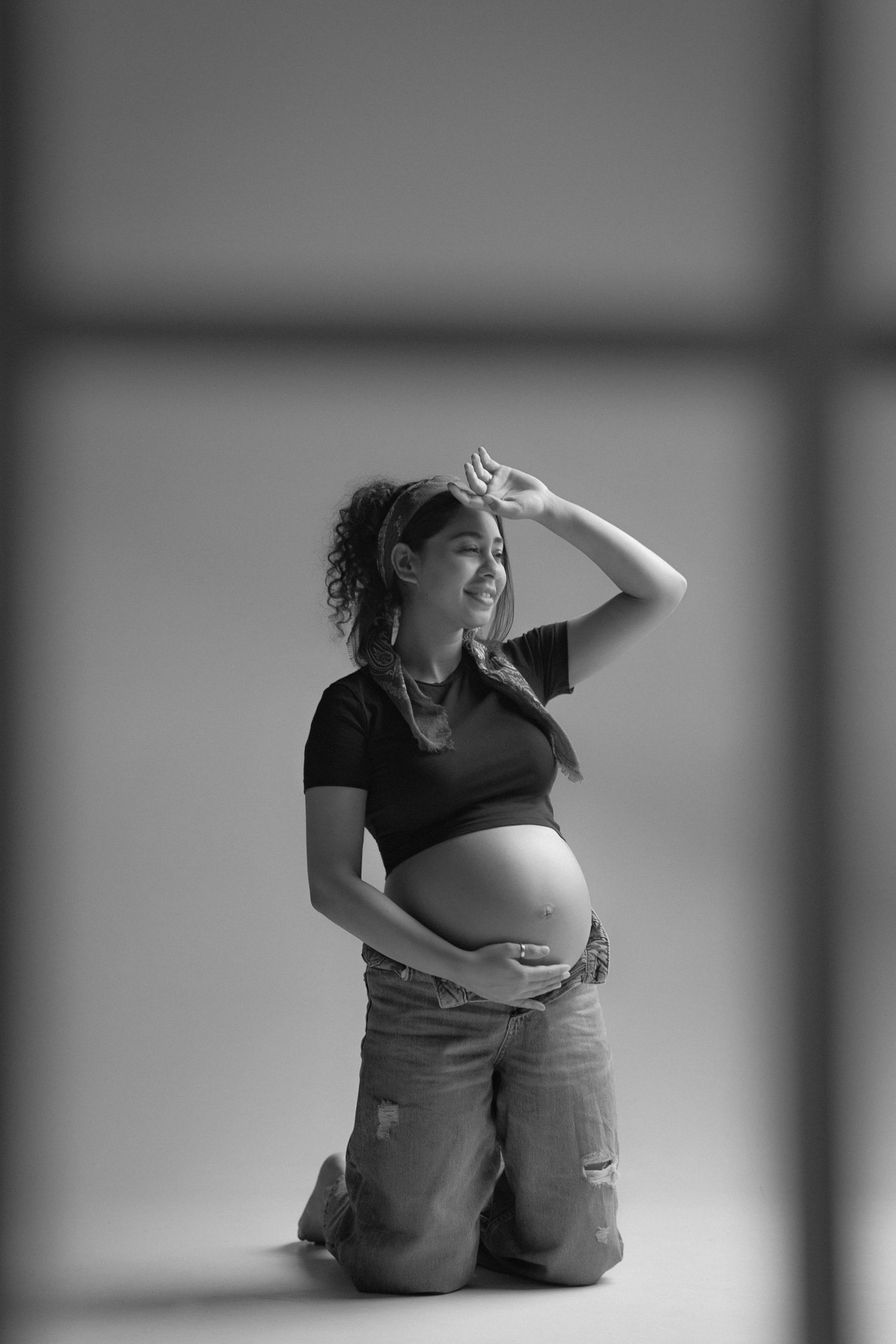 Black and white photo of a smiling pregnant woman kneeling, cradling her baby bump in ripped jeans and a black crop top.