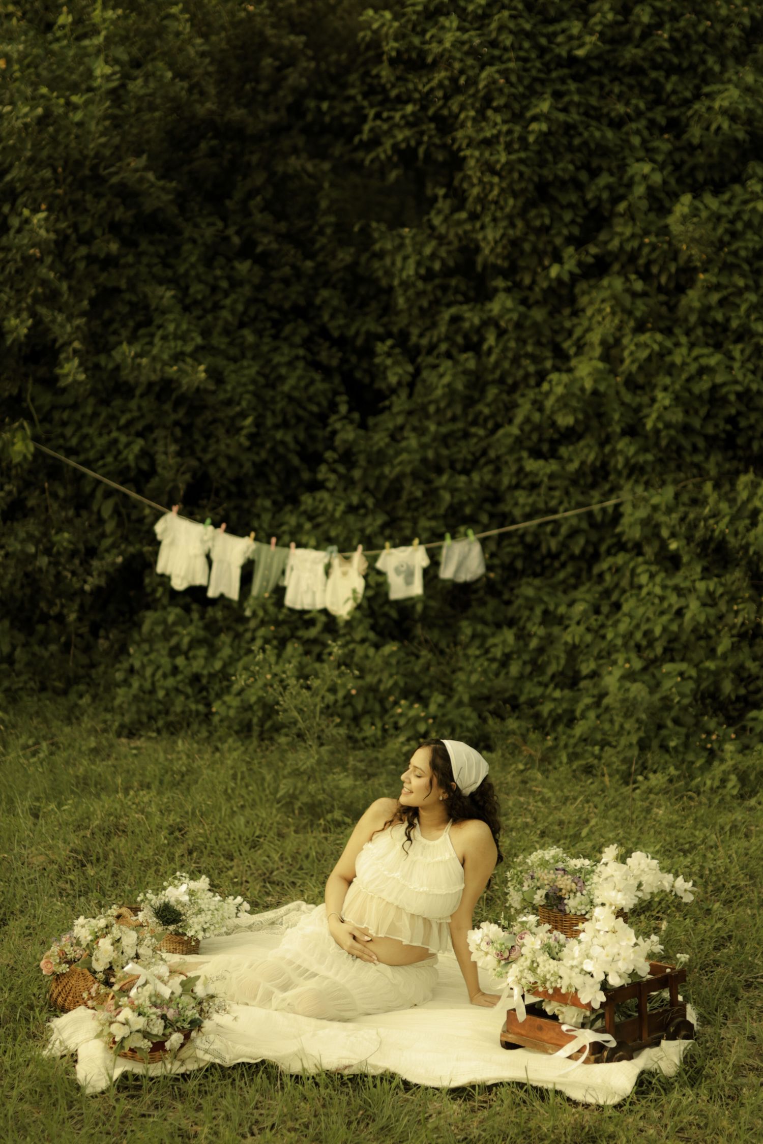 Pregnant woman in white dress sitting on blanket outdoors with flowers, baby clothes hanging on line behind her.