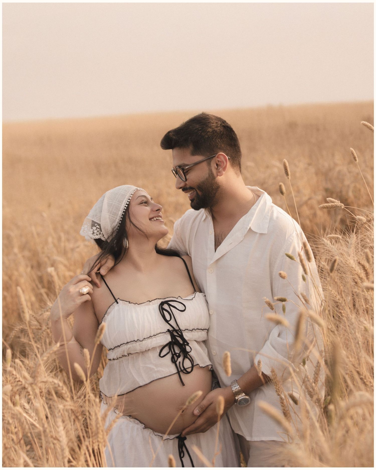 Pregnant couple in white outfits sharing a tender moment in a golden wheat field during a maternity photoshoot.