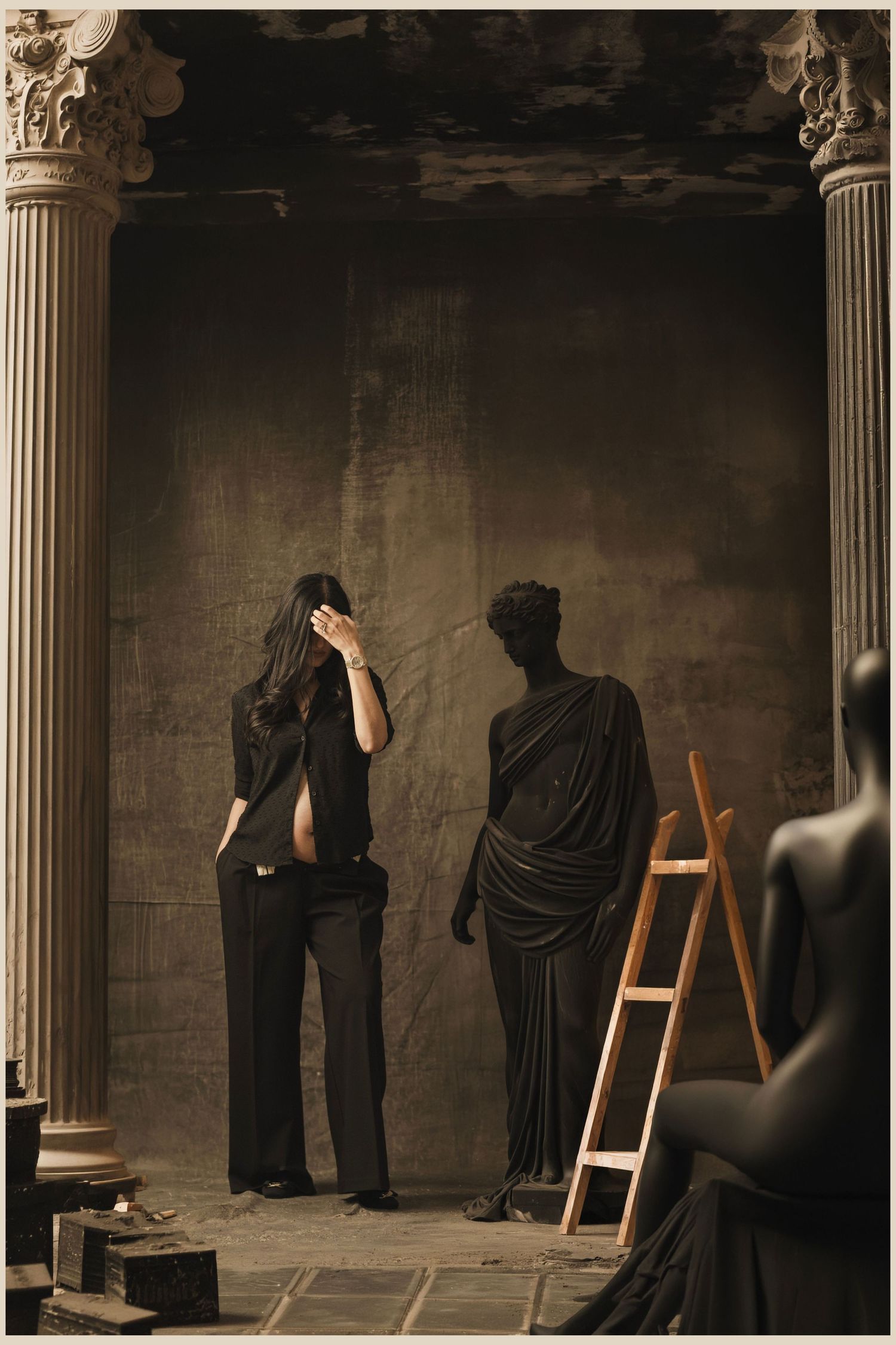 A figure in black stands before a dark classical backdrop with columns, a draped statue, and an easel.