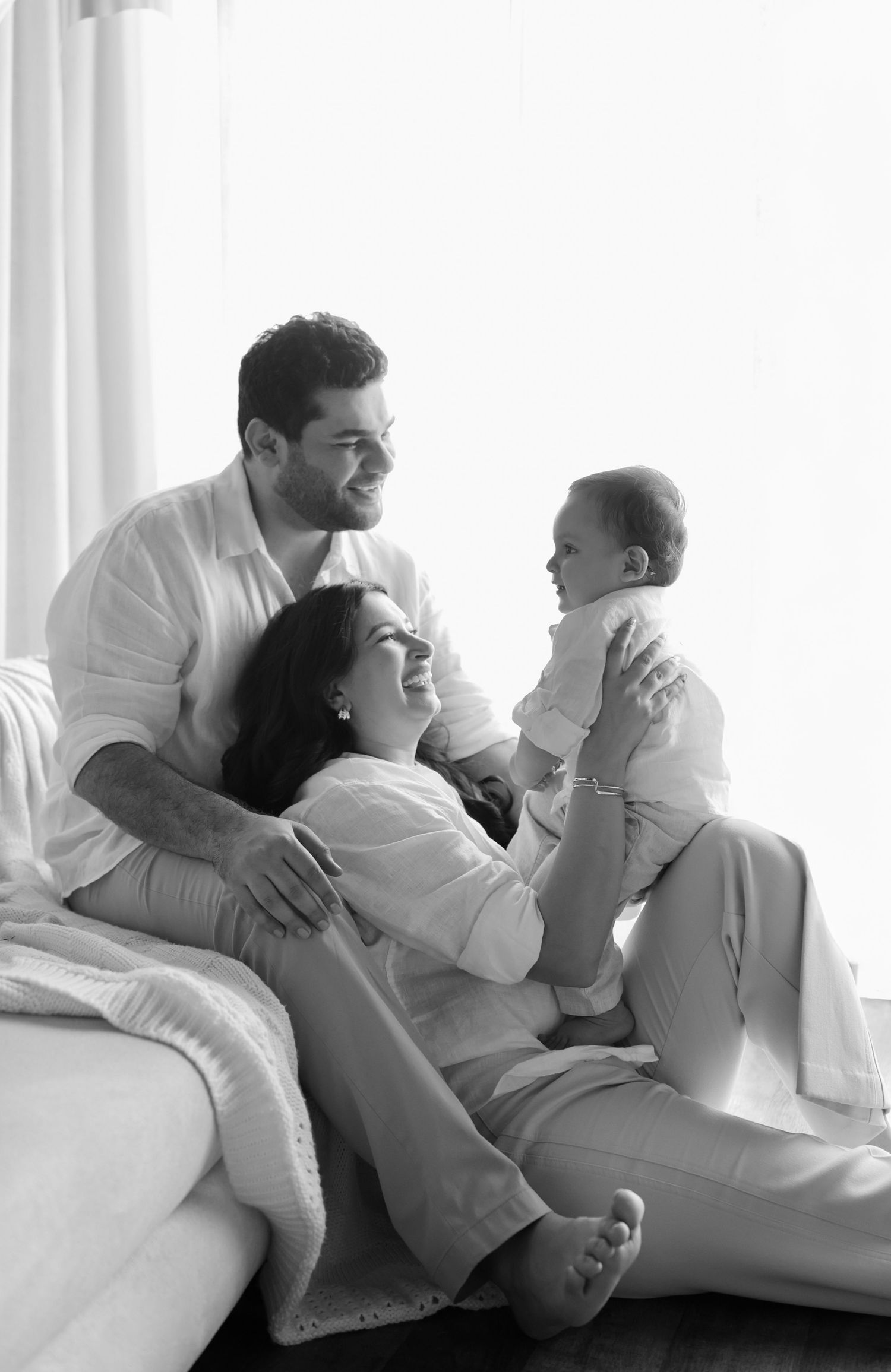Black and white photo of happy parents holding their smiling baby together near a bright window at home.