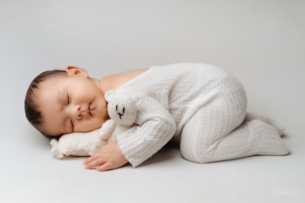 Sleeping newborn baby in white knit onesie cuddling a small stuffed animal toy on a light gray background.