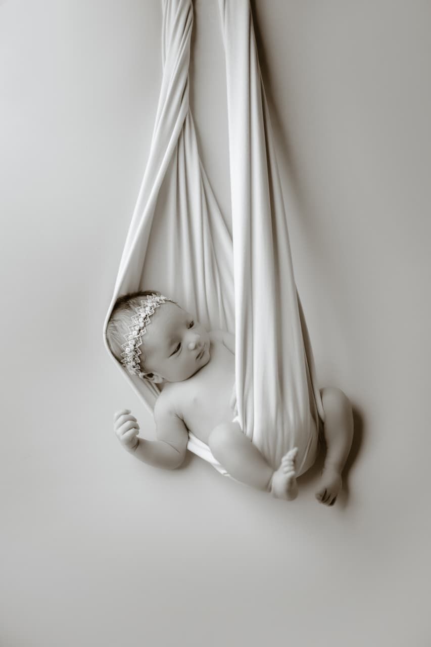 Newborn baby girl sleeping peacefully in a white fabric hammock swing, wearing a delicate lace headband.