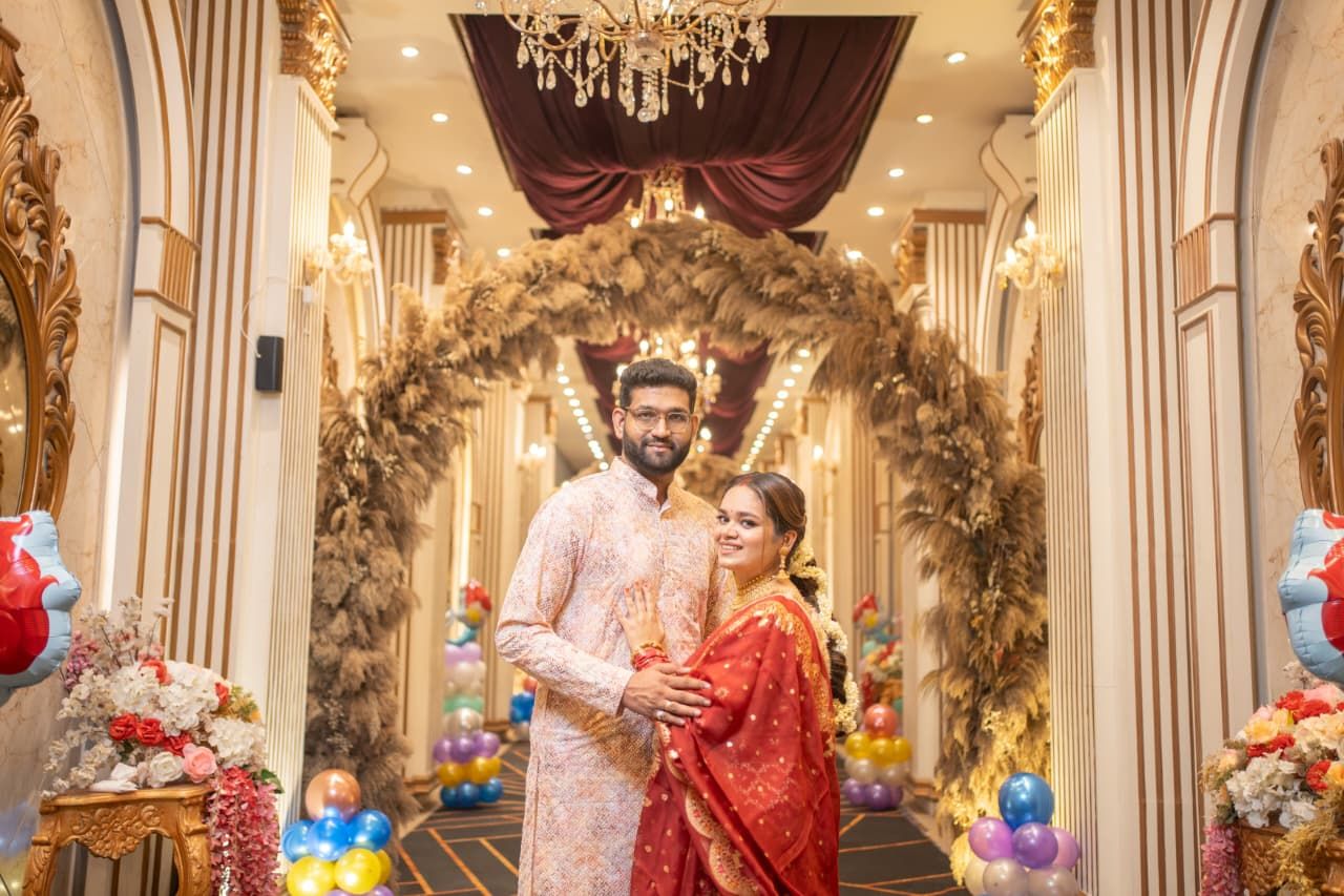 Couple in traditional South Asian attire posing under floral arch in elegant decorated wedding venue.