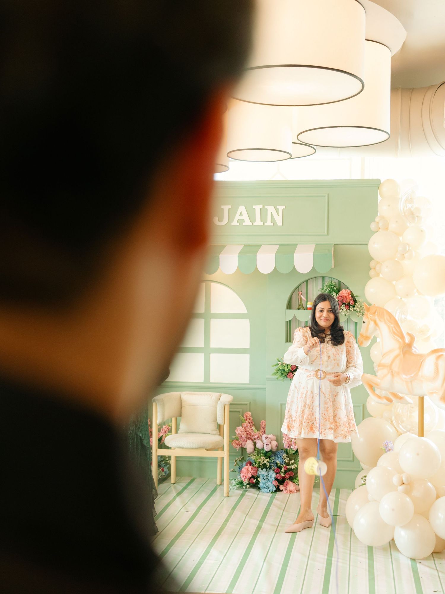Woman in floral dress poses at elegant mint green JAIN backdrop with balloons and flowers at a celebration event.
