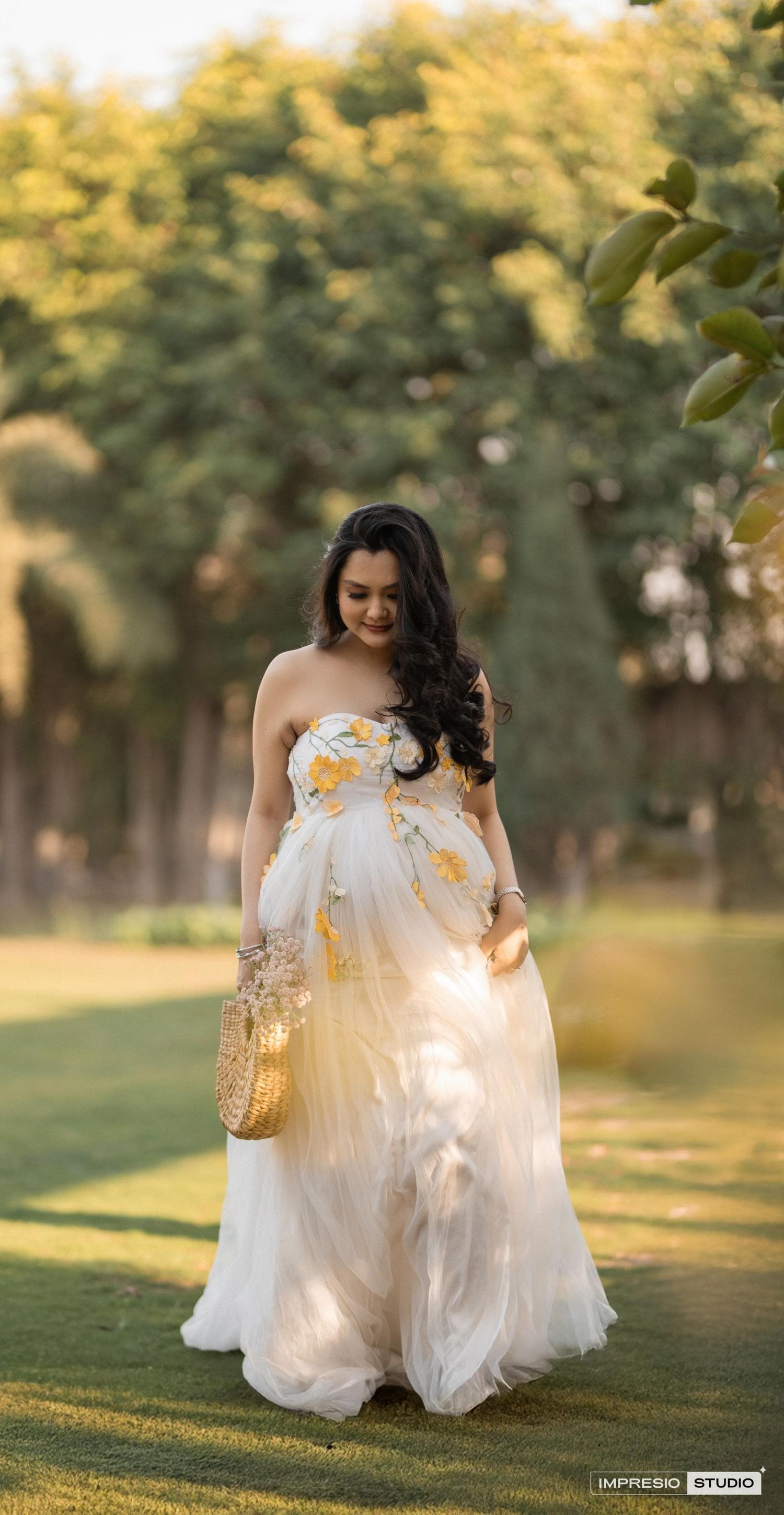 A flowing white and gold maternity gown catches the evening light in an outdoor garden setting.
