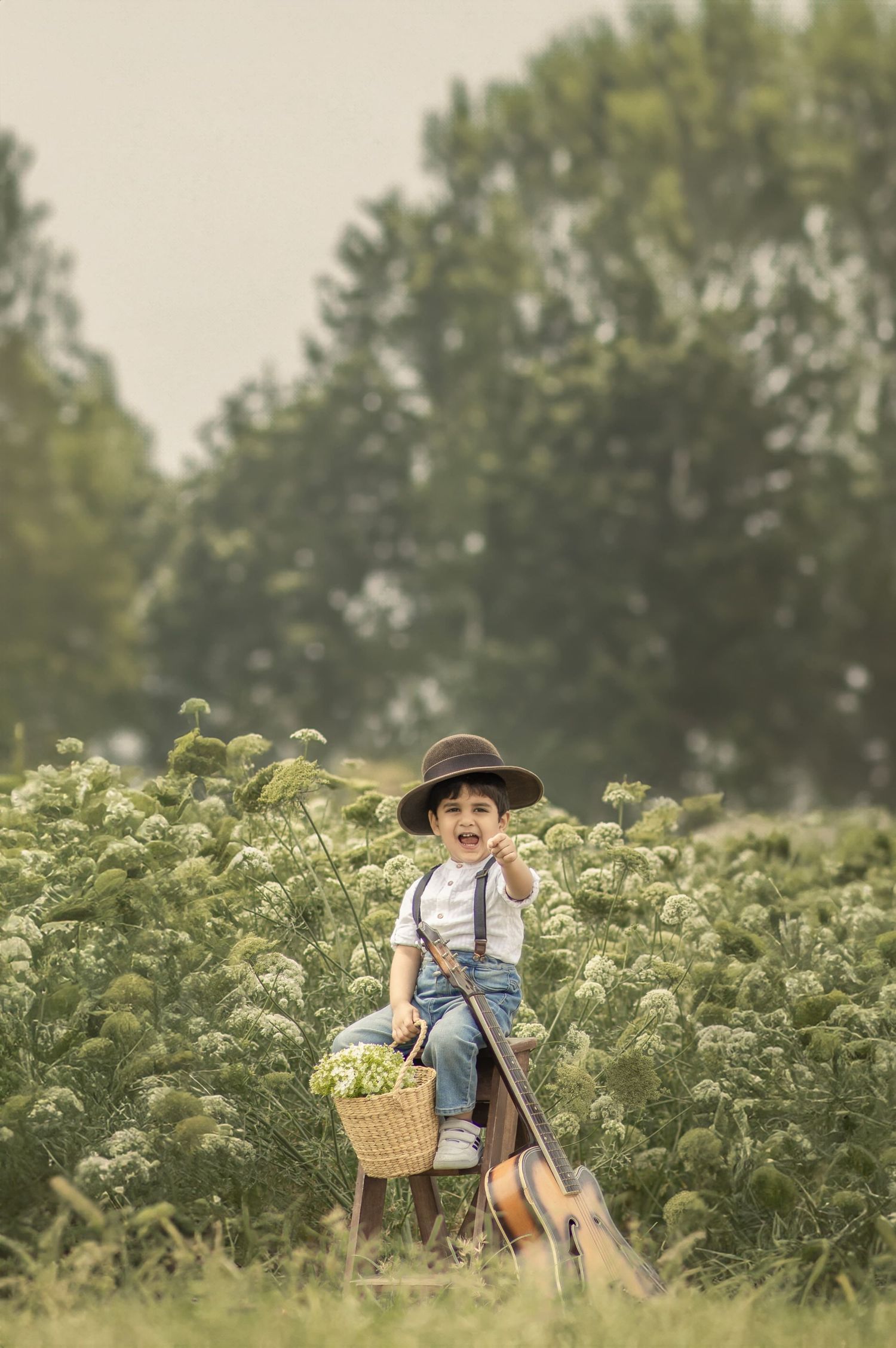 Child in countryside wearing hat and suspenders while carrying flower basket among tall plants for pre birthday photoshoot