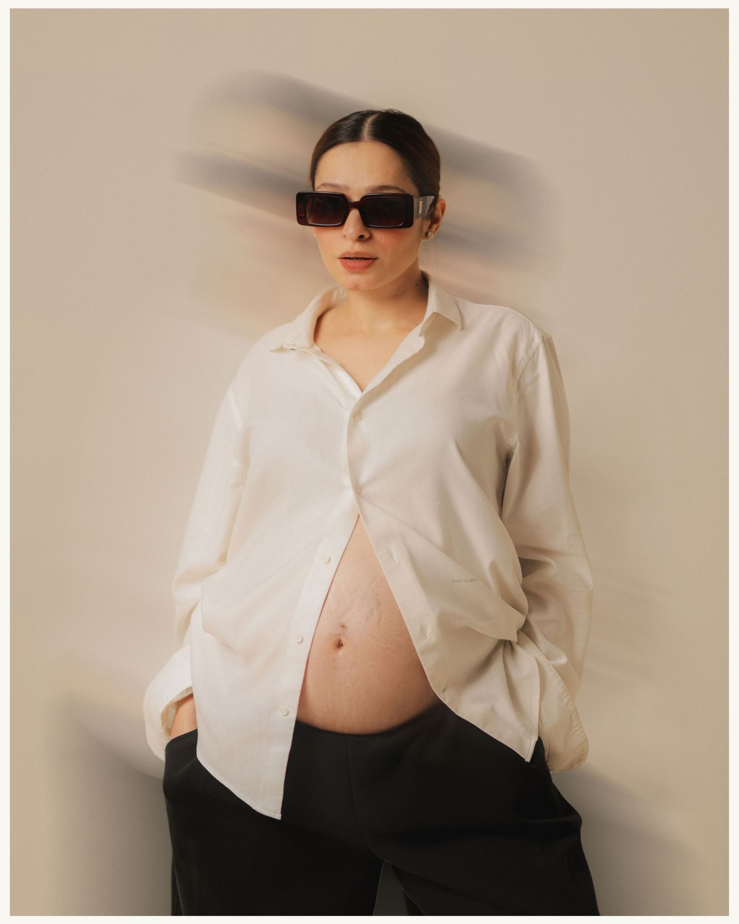 Pregnant woman in white oversized shirt and black sunglasses posing with baby bump exposed against beige background.