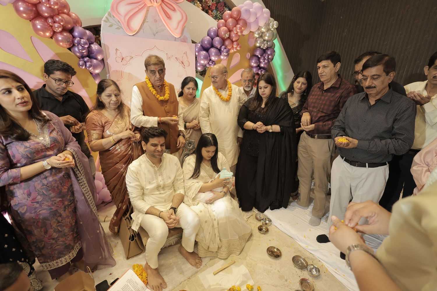 Family and guests in traditional Indian attire gather around a couple holding a newborn during a ceremony decorated with pink and purple bal