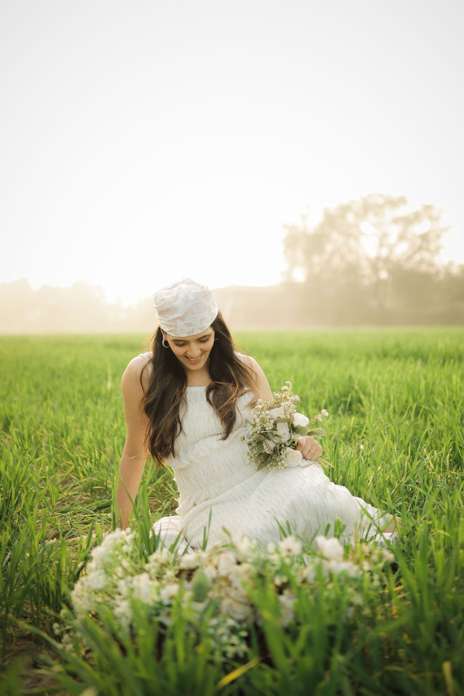 A bride in a white dress and cap sits in a sunlit meadow holding a bouquet of wildflowers at golden hour.